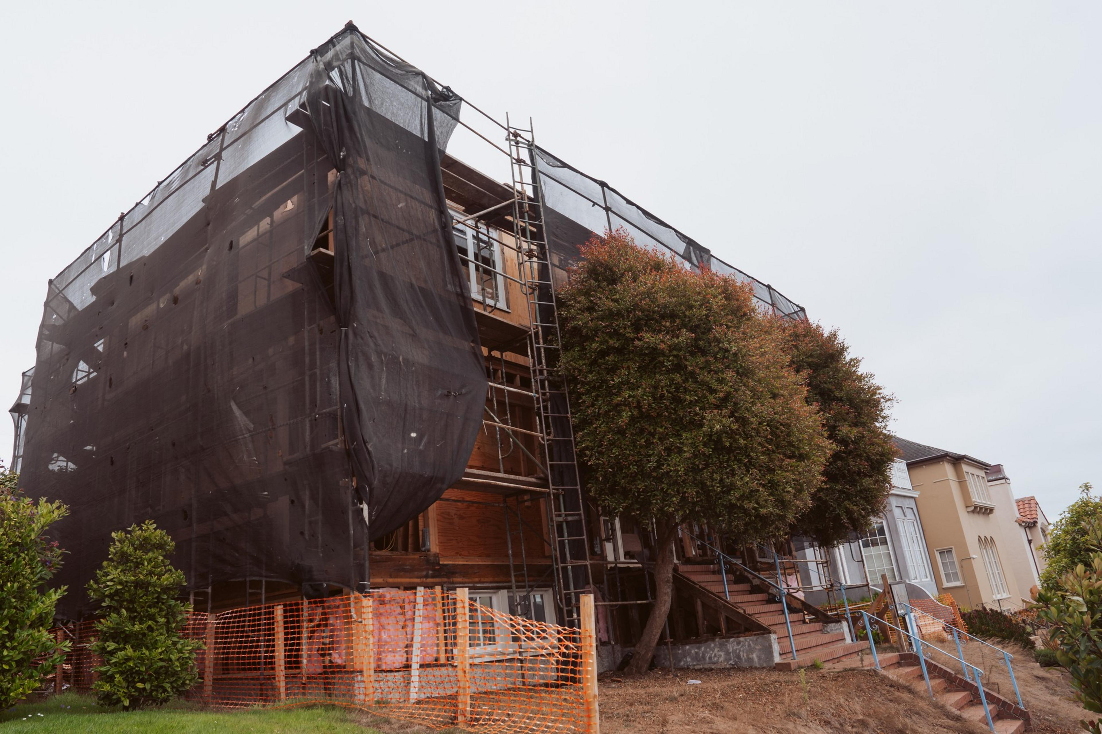 A building under construction with scaffolding and a black protective mesh, beside trees and a fenced-off area.