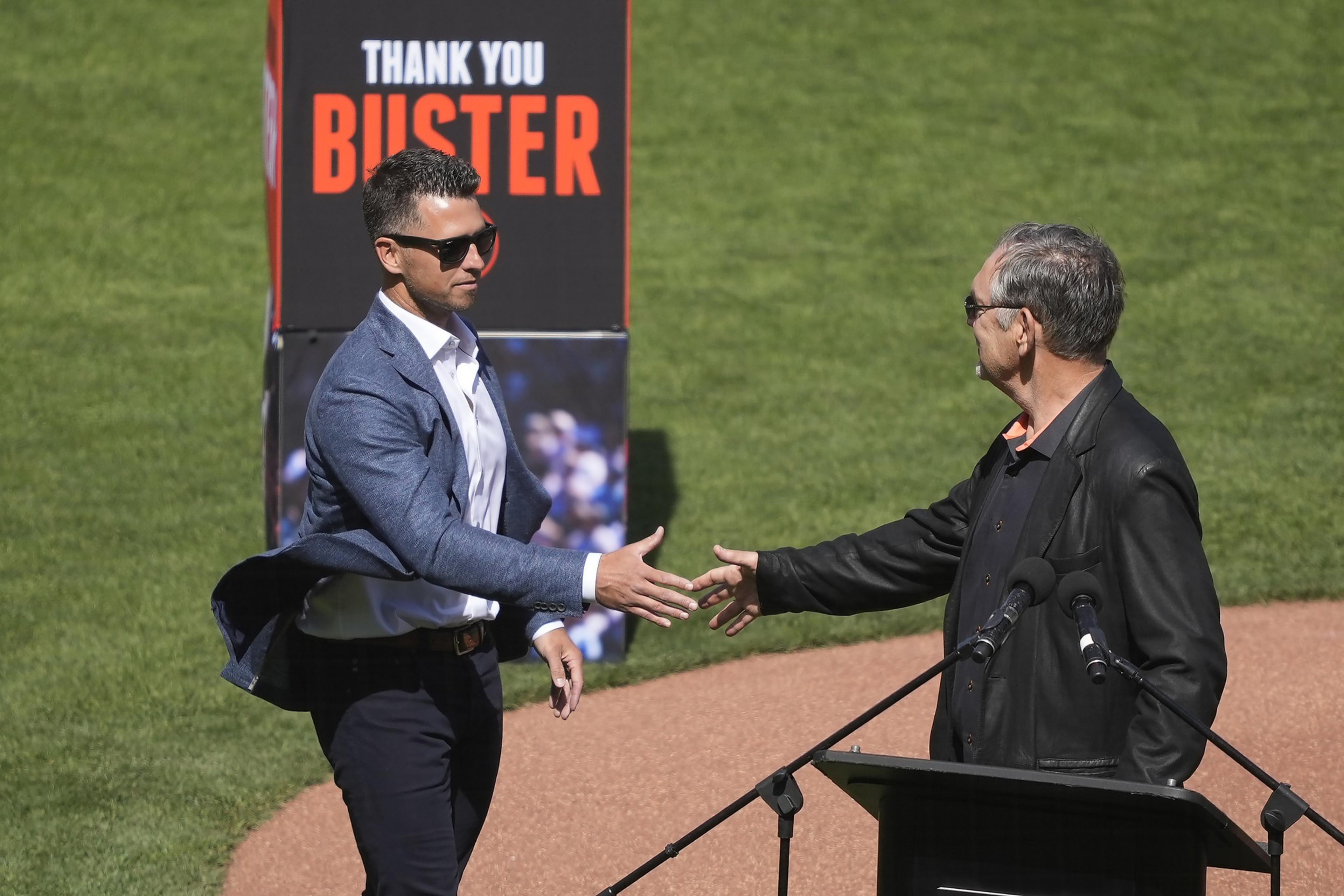 Two men, both wearing sunglasses, shake hands on a baseball field. One wears a blue blazer, the other a black jacket. A sign says &quot;Thank You Buster.&quot;
