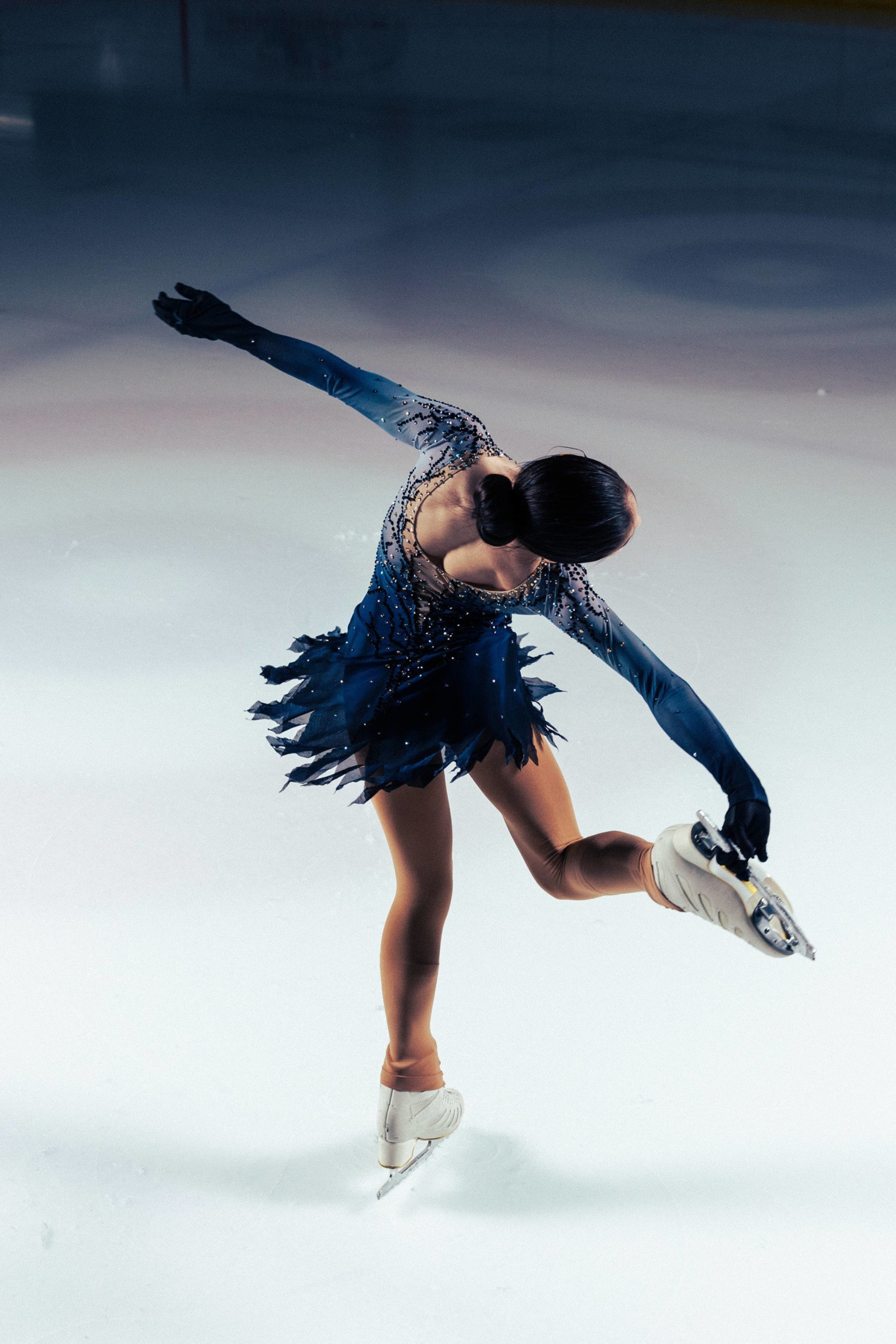 A figure skater in a dark blue, sparkly dress spins on one leg, holding her other leg behind her on an ice rink.