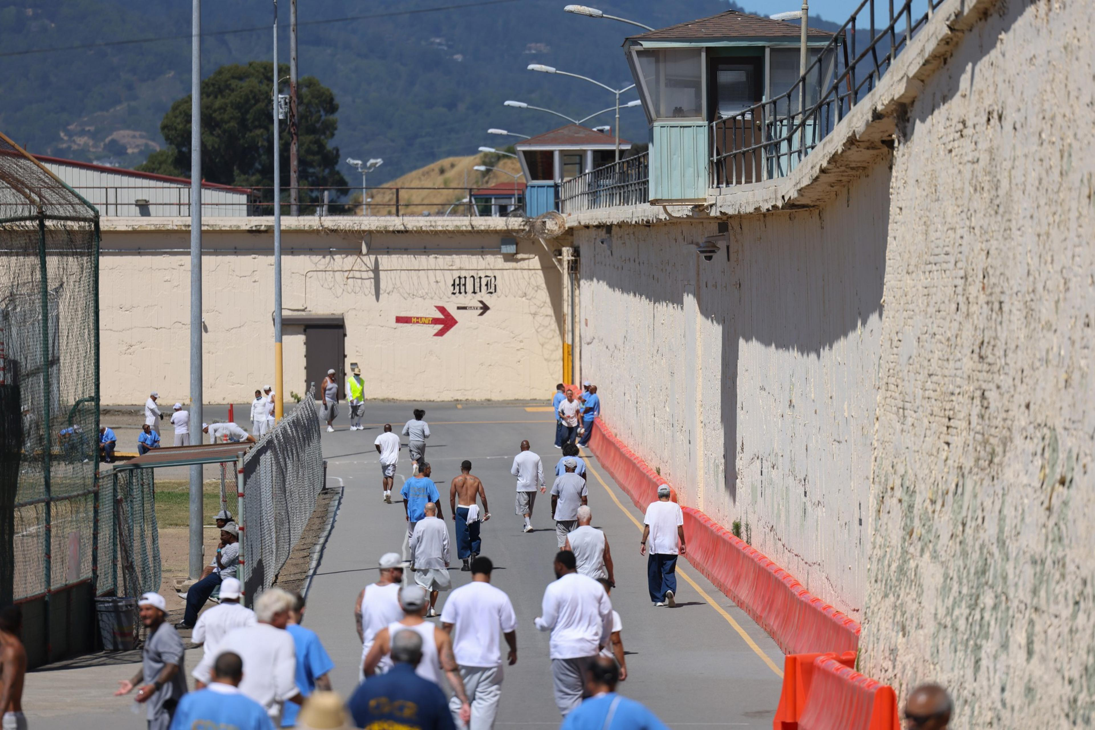 Inmates walk in a yard by a high wall with a guard tower above. The mood seems orderly.