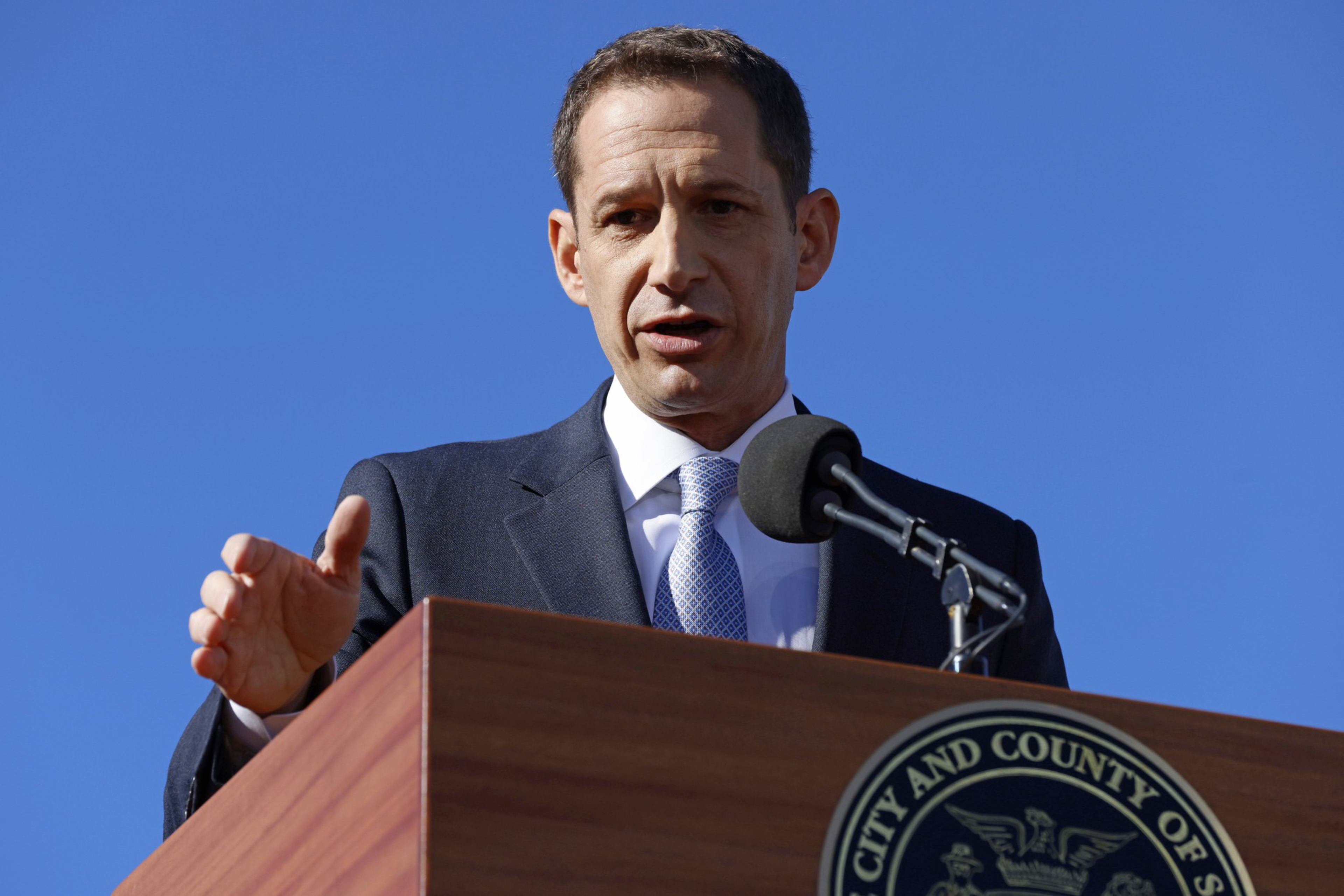 A person in a suit speaks at a podium with a microphone in front of a clear blue sky. The podium has an insignia for the &quot;City and County of&quot; an unspecified place.