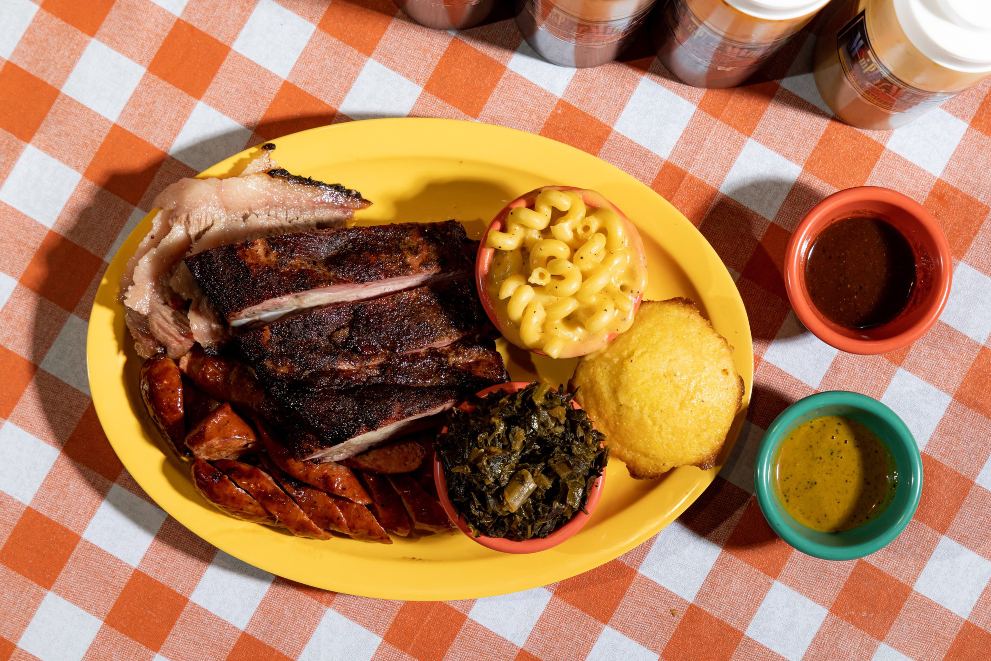 Barbecue plate with ribs, sausage, sliced meat, macaroni and cheese, cooked greens, cornbread, and two dipping sauces on a yellow plate.