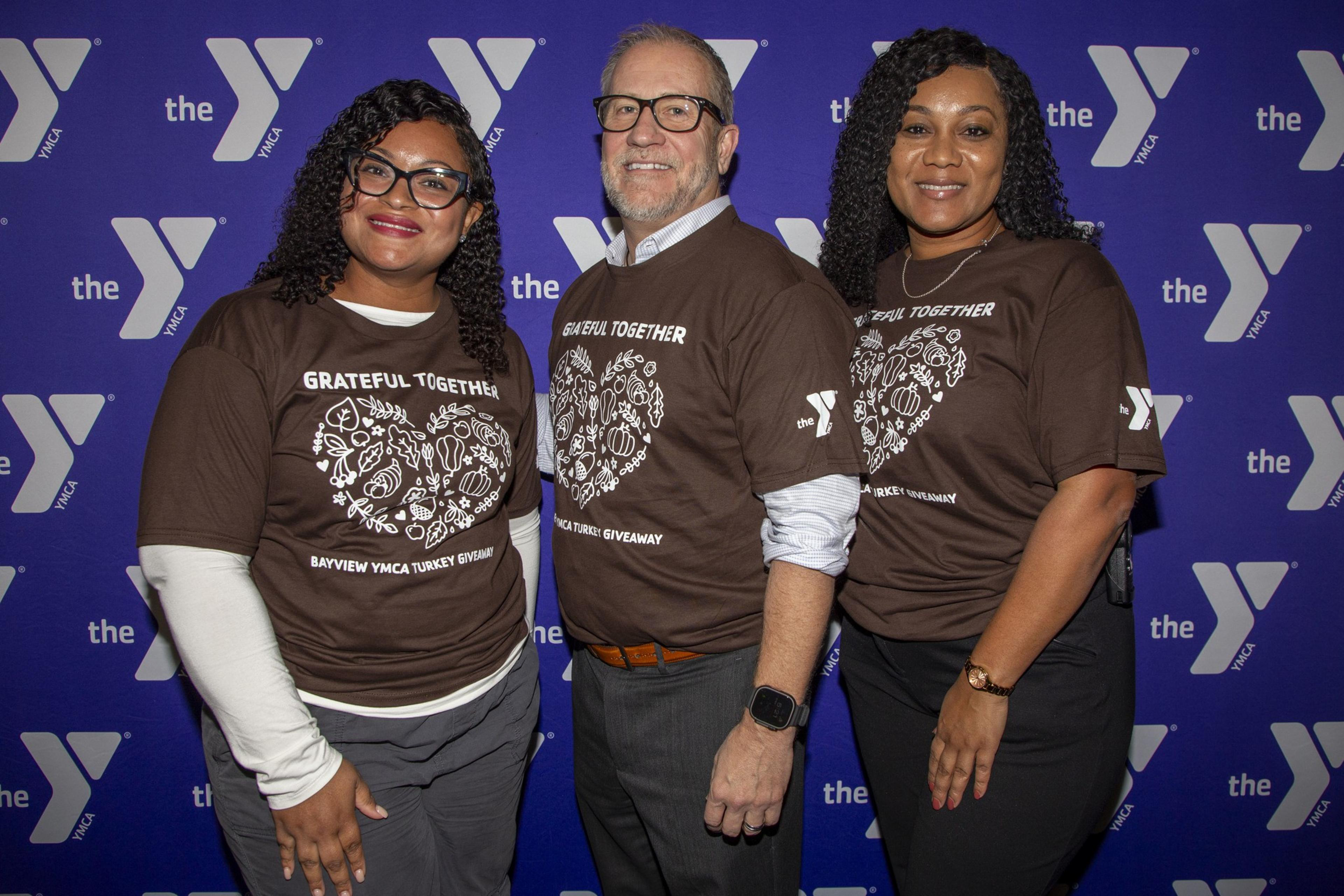 Three people smile at the camera, wearing matching brown shirts with &quot;Grateful Together&quot; on them, in front of a YMCA backdrop.