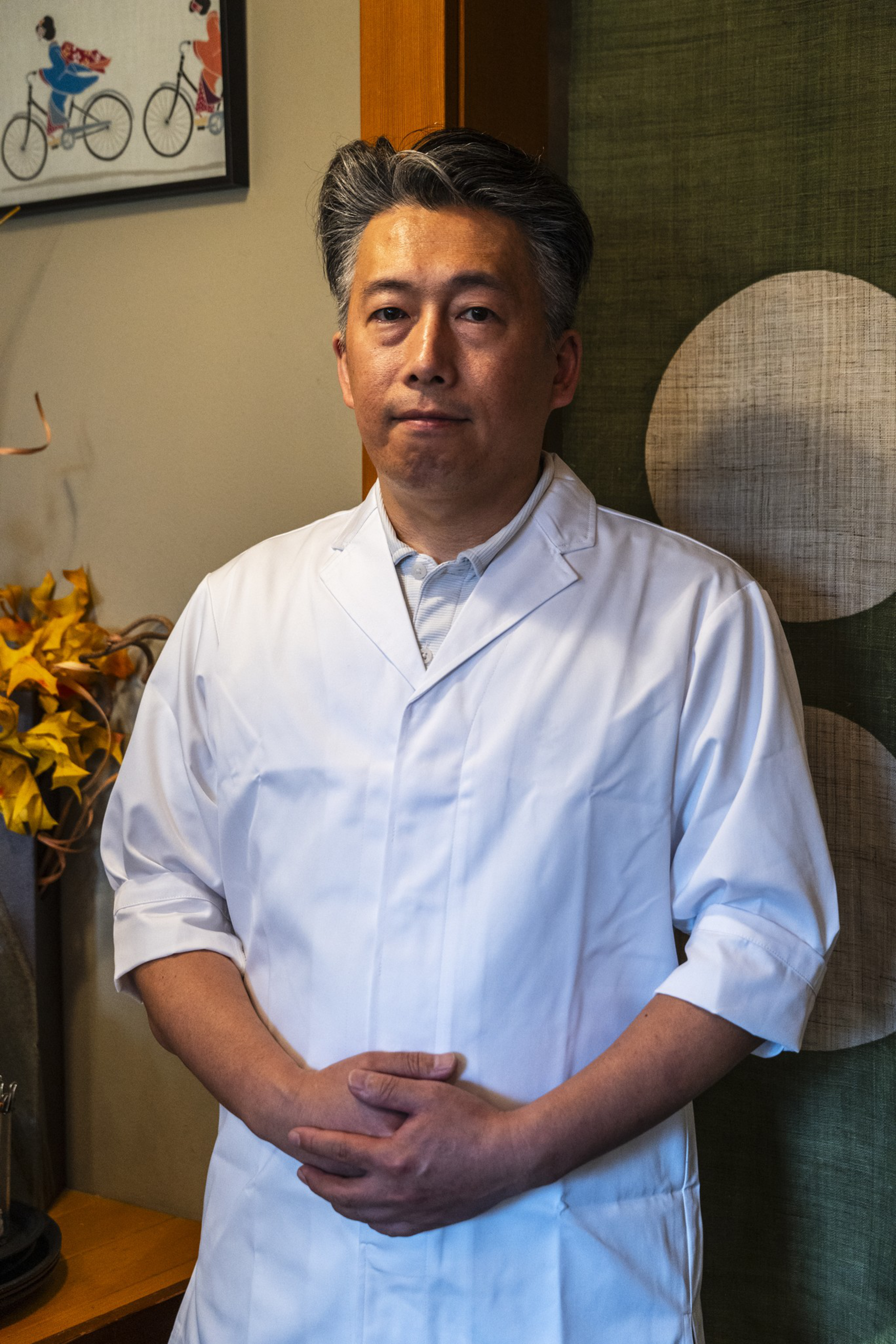 A man with salt-and-pepper hair stands with hands clasped, wearing a white coat, in a room with green and beige wall decor.