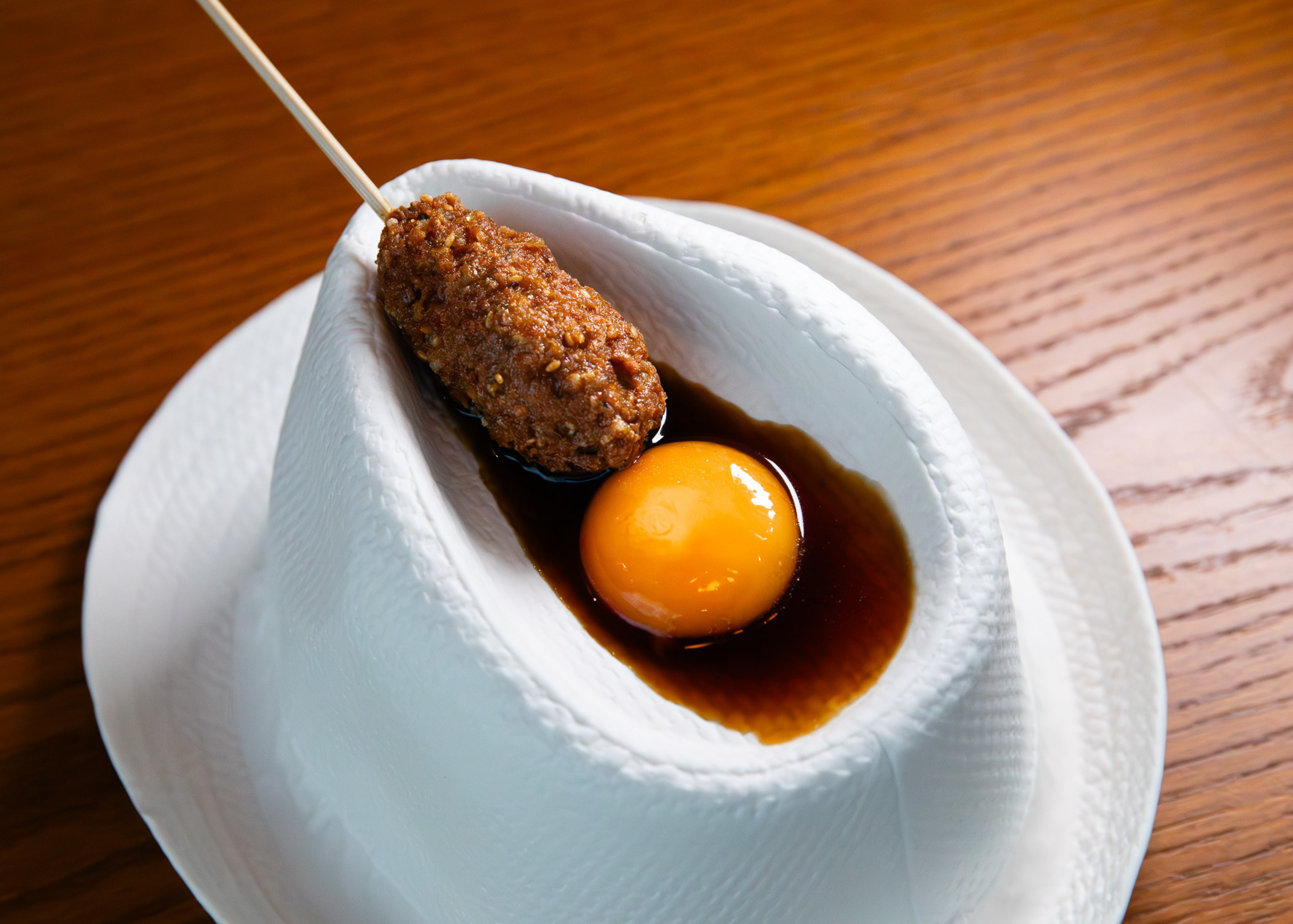 A deep-fried breaded skewer and a raw egg yolk sit in a small pool of dark sauce on a white napkin, placed on a white plate over a wooden table.