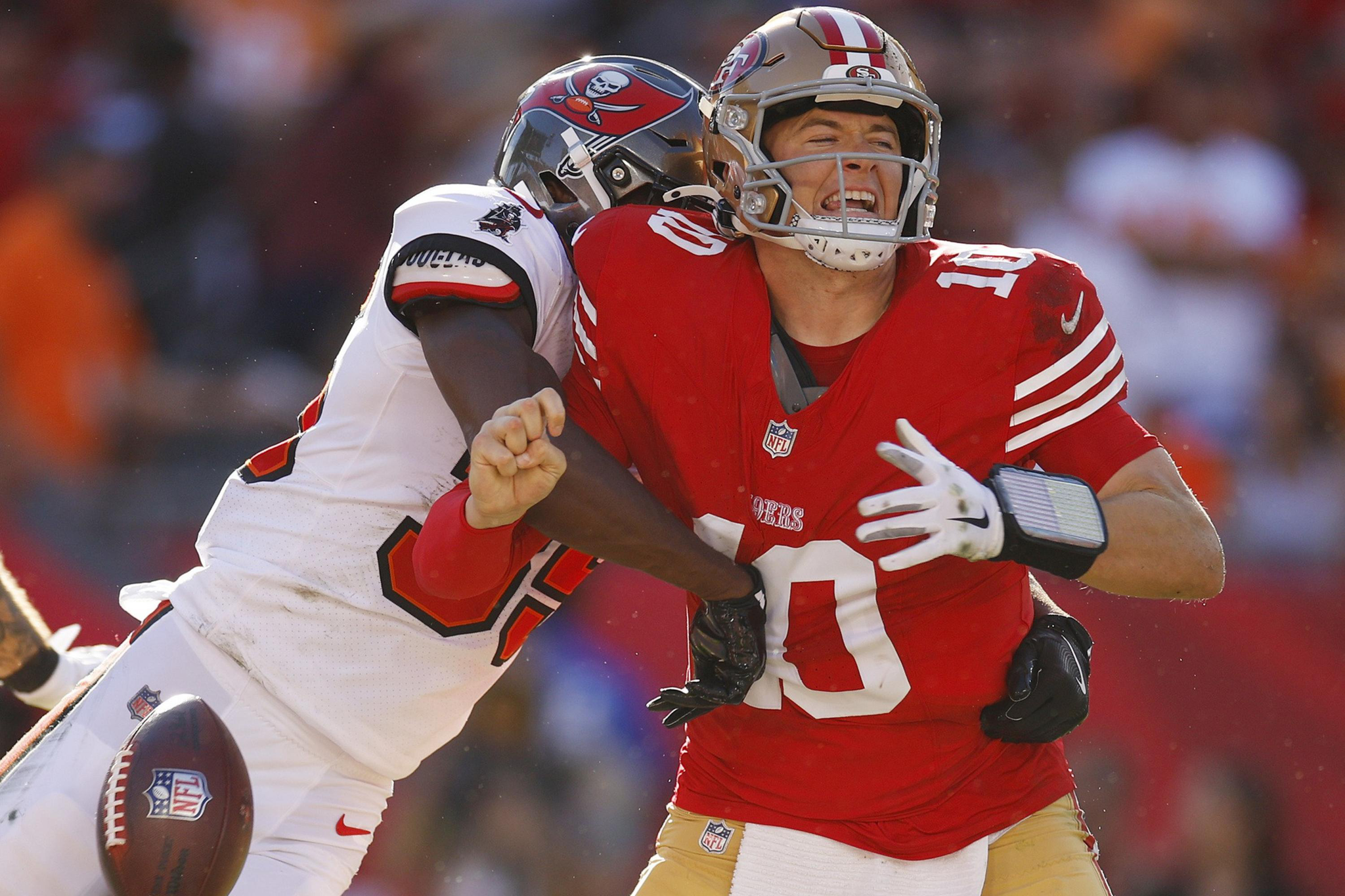 A football player in a white Tampa Bay Buccaneers jersey tackles a San Francisco 49ers quarterback in a red jersey, causing the ball to come loose.