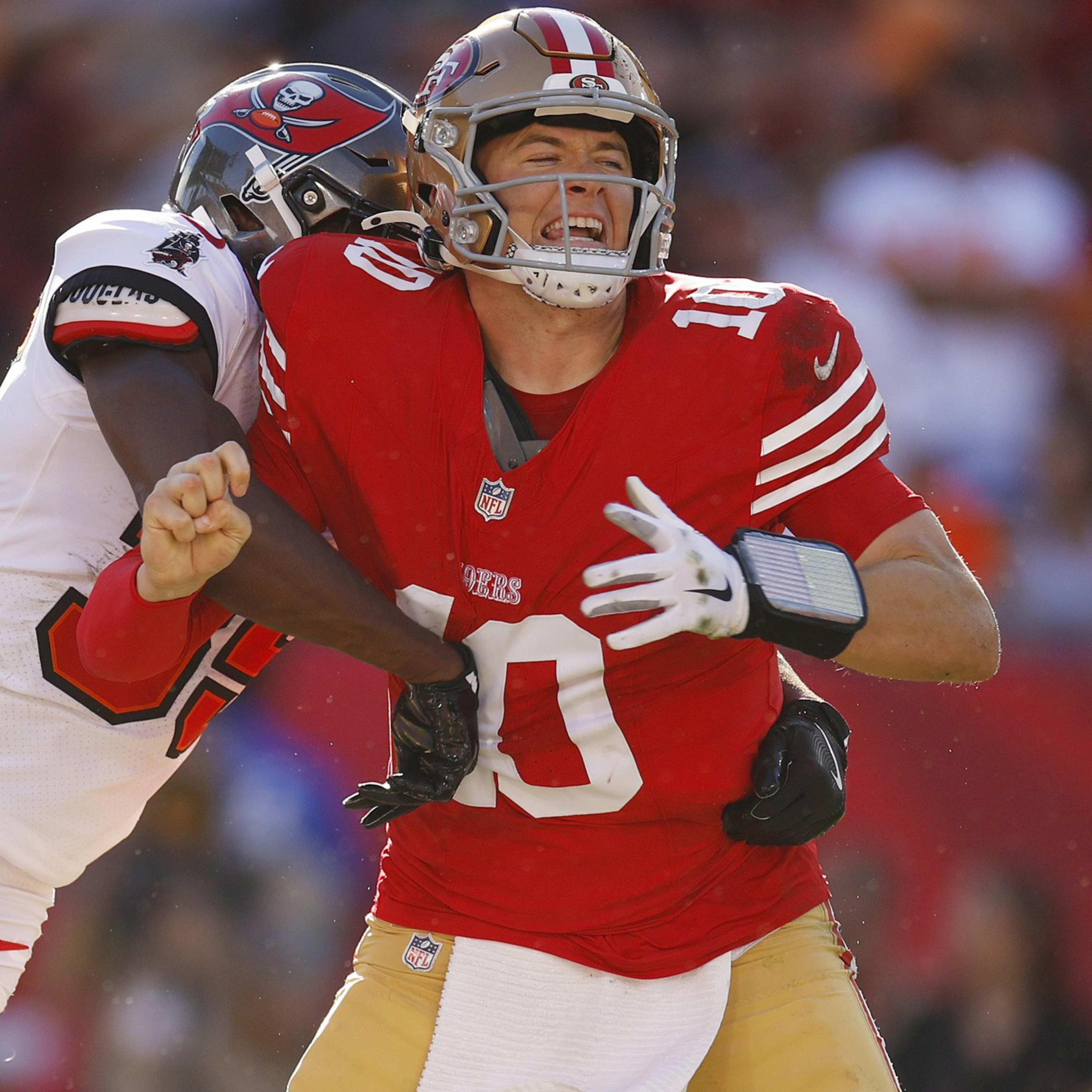 A football player in a white Tampa Bay Buccaneers jersey tackles a San Francisco 49ers quarterback in a red jersey, causing the ball to come loose.