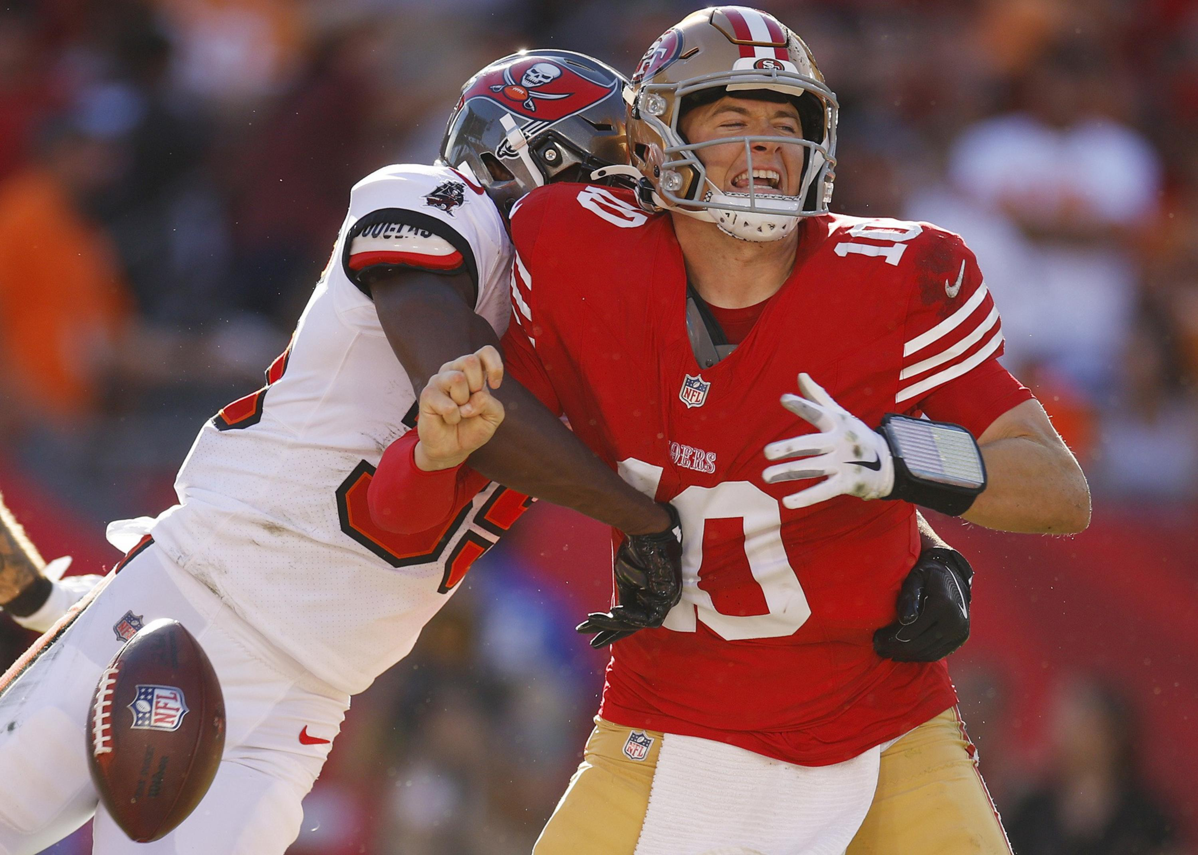 A football player in a white Tampa Bay Buccaneers jersey tackles a San Francisco 49ers quarterback in a red jersey, causing the ball to come loose.