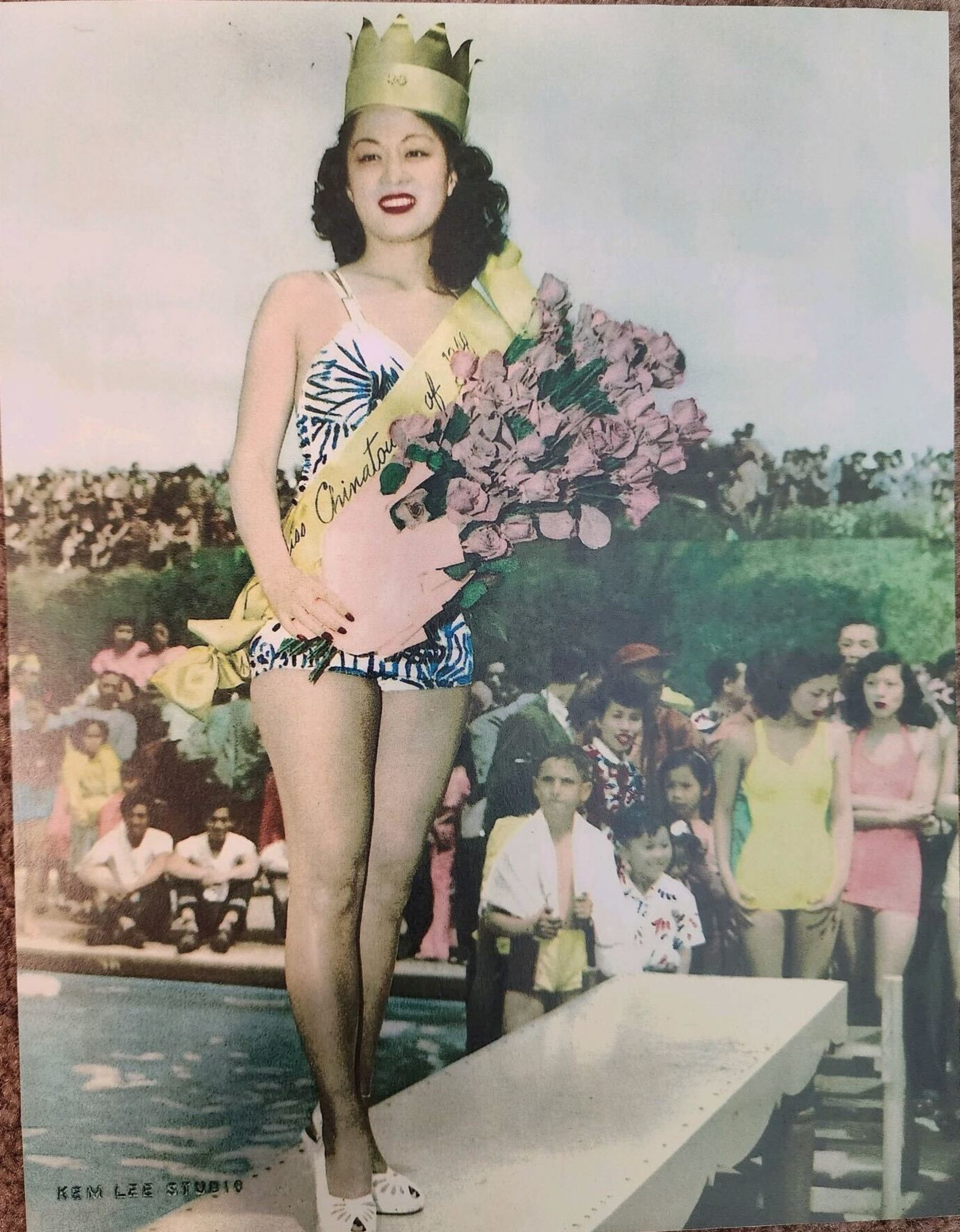 A woman in a floral swimsuit, crown, and sash holds a bouquet of flowers and smiles, standing on a platform by a pool with a crowd behind her.