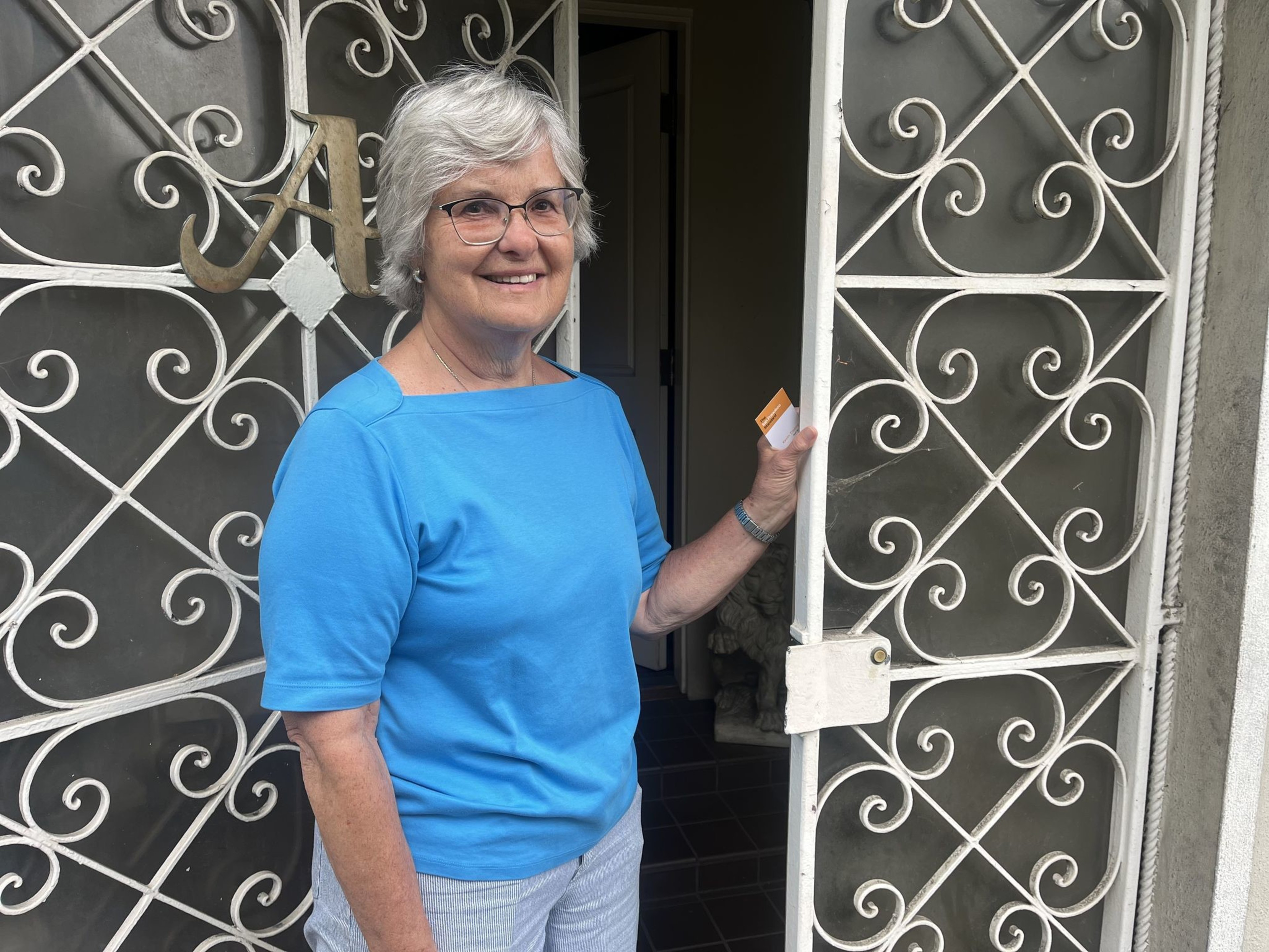A smiling woman with gray hair, wearing a blue shirt, holds a card at an ornate metal gate.