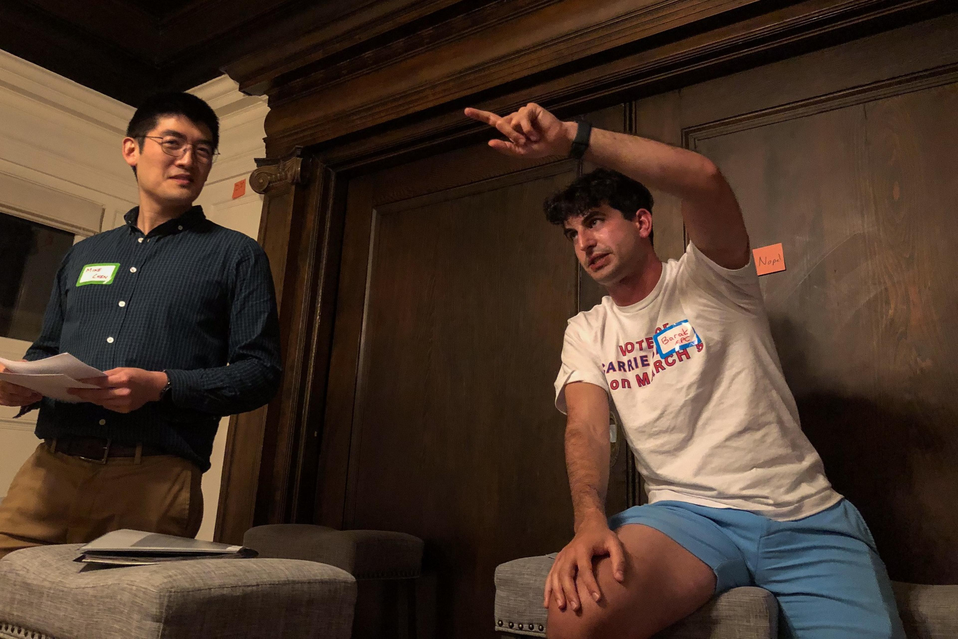 Two men in a room, one standing with papers, the other sitting and pointing, both look engaged in a discussion.