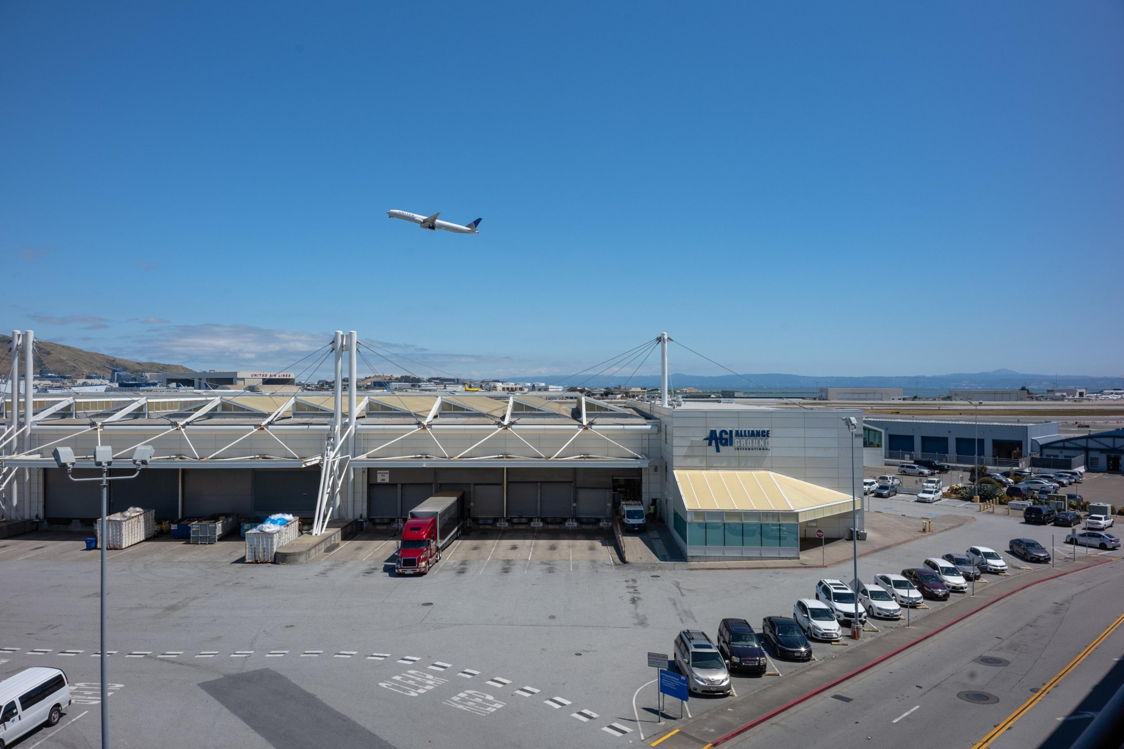 An airplane is flying over a warehouse area with &quot;AGI Alliance Ground&quot; signage. Below are parked cars, shipping containers, and a red semi-truck. The sky is clear.