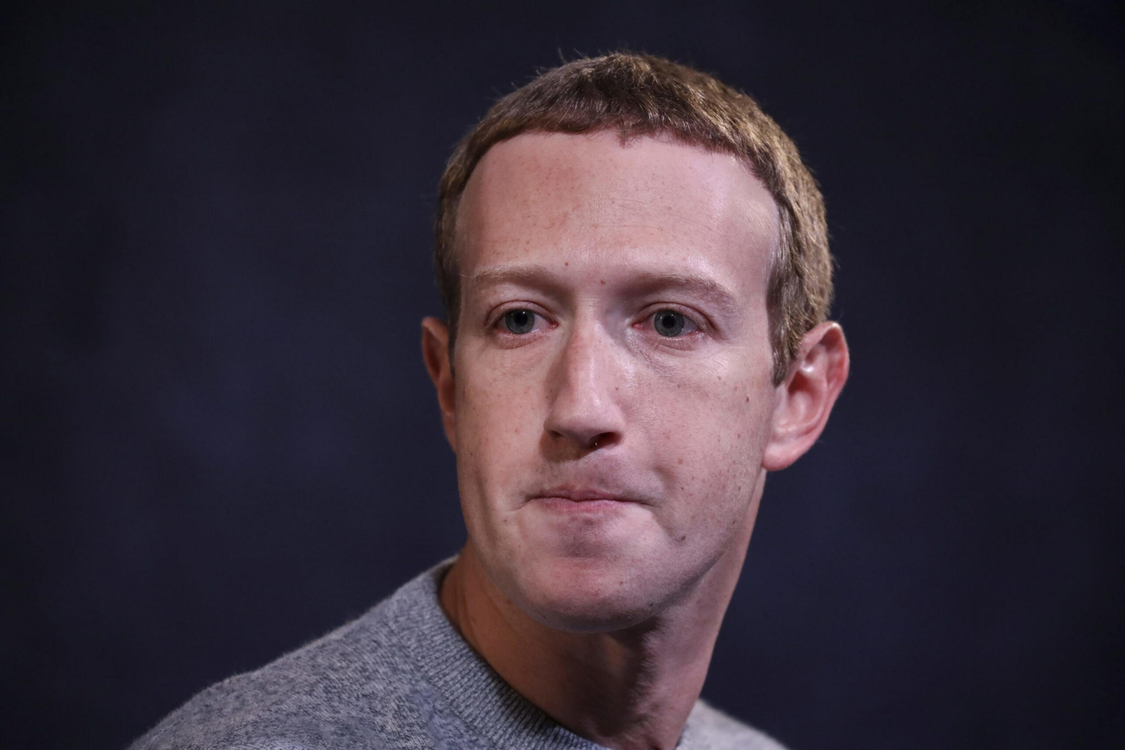 A man with short light brown hair and pale skin looks slightly to the side, wearing a gray sweater against a dark, blurred background.