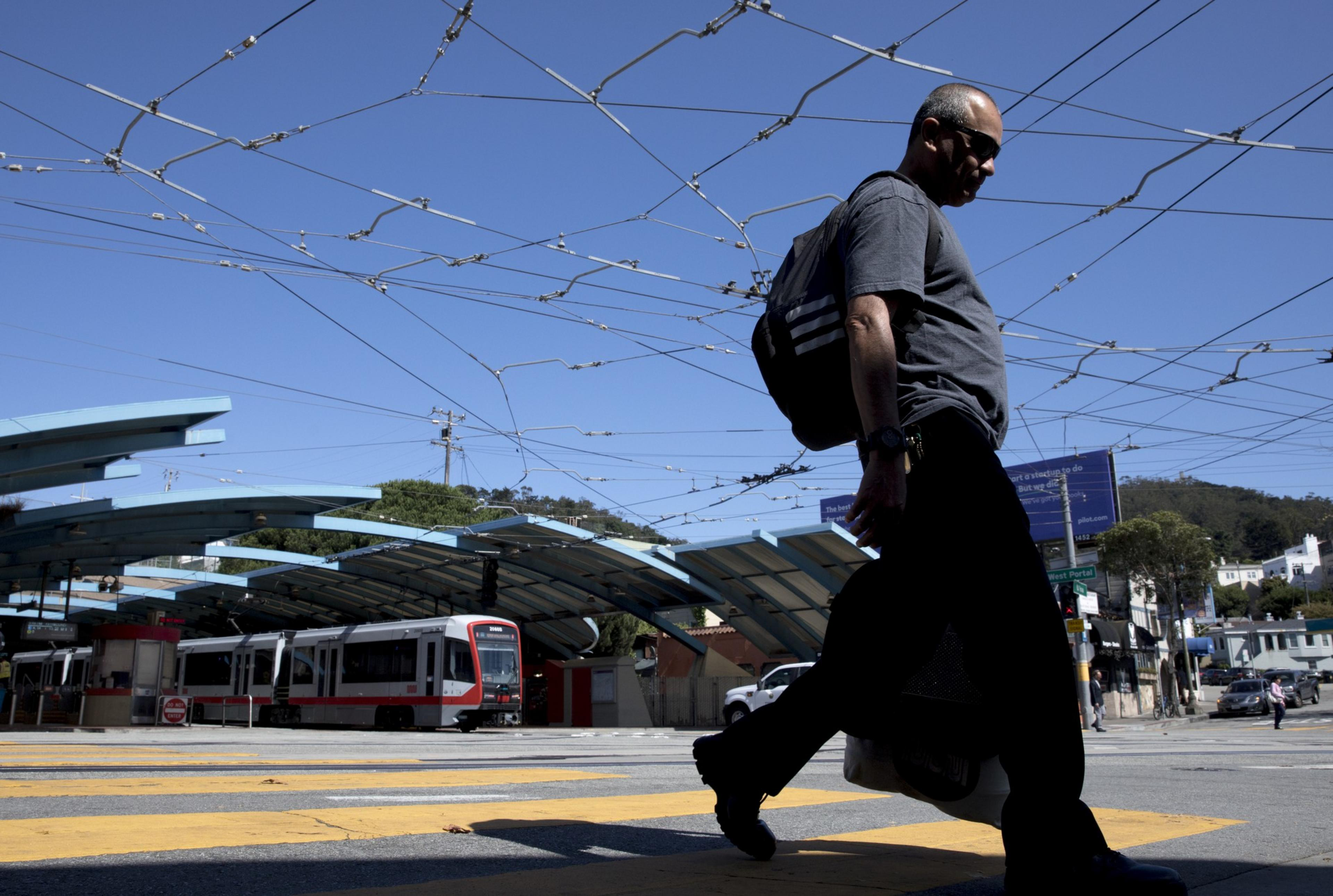 A man walks across a street with a tram station and overhead wires in the background.