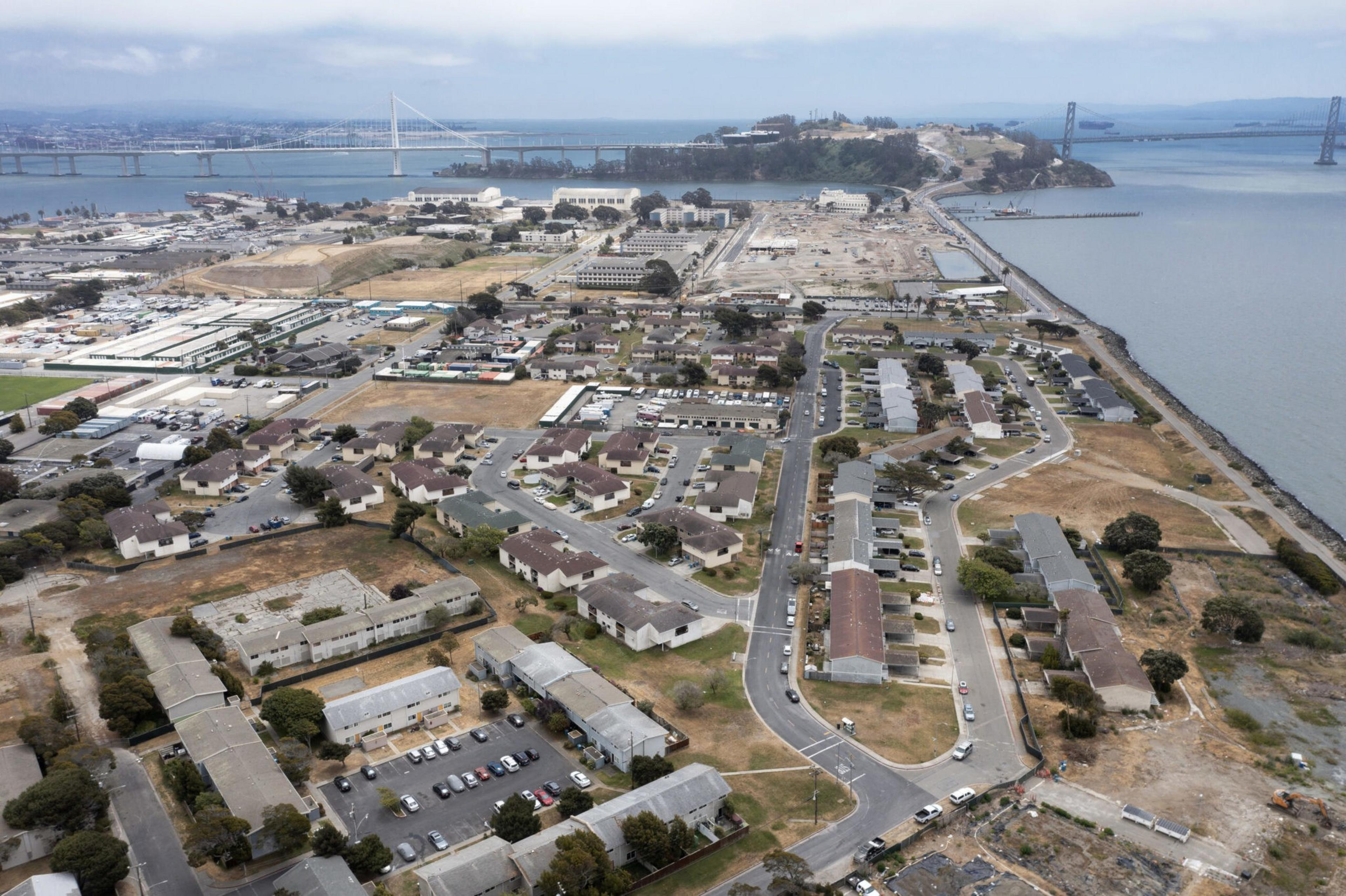 The image shows an aerial view of a peninsula with residential housing, roads, and some industrial areas. Two bridges span the water in the background.