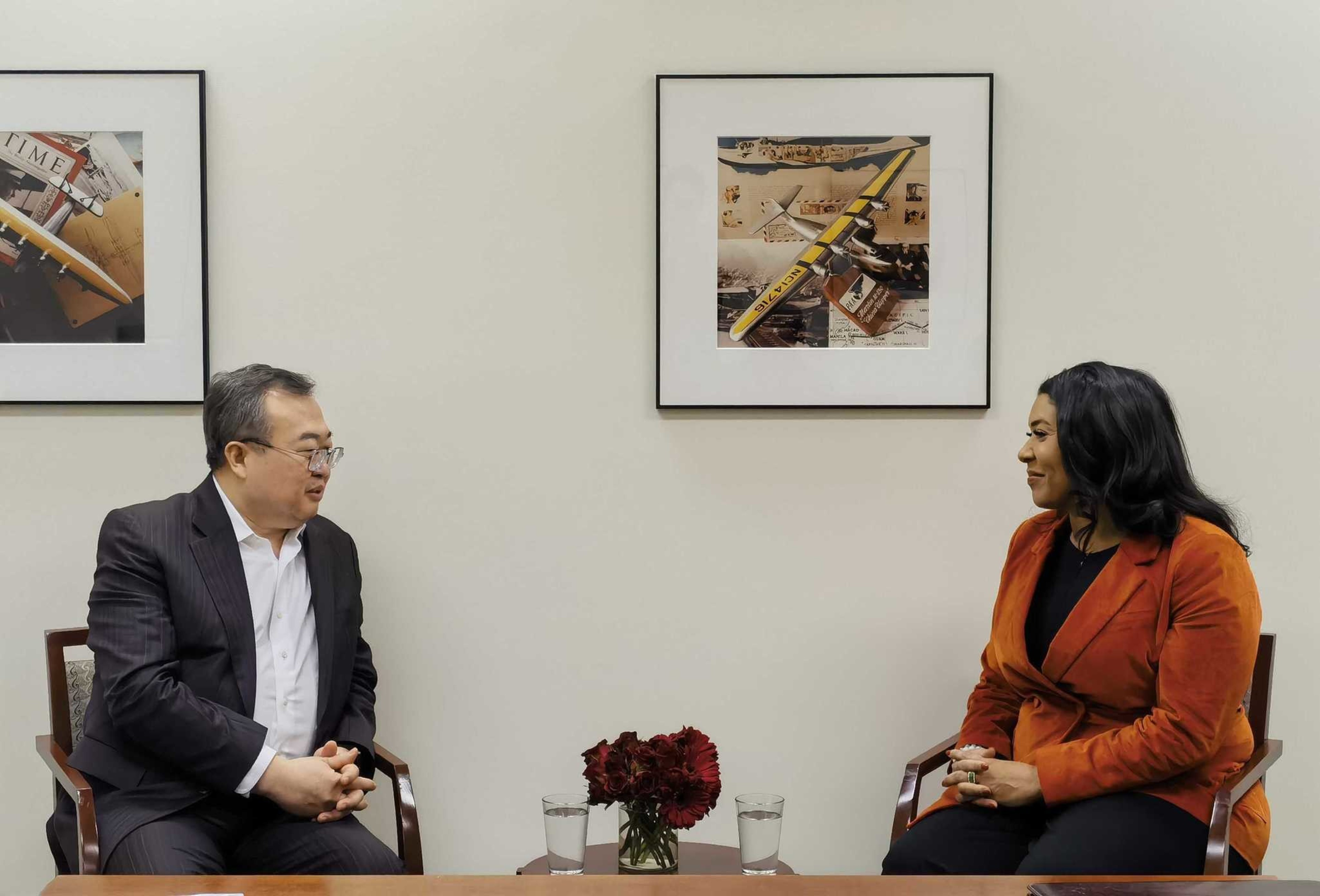 Two people sit facing each other, smiling, with framed photos on the wall behind them and a bouquet between them.