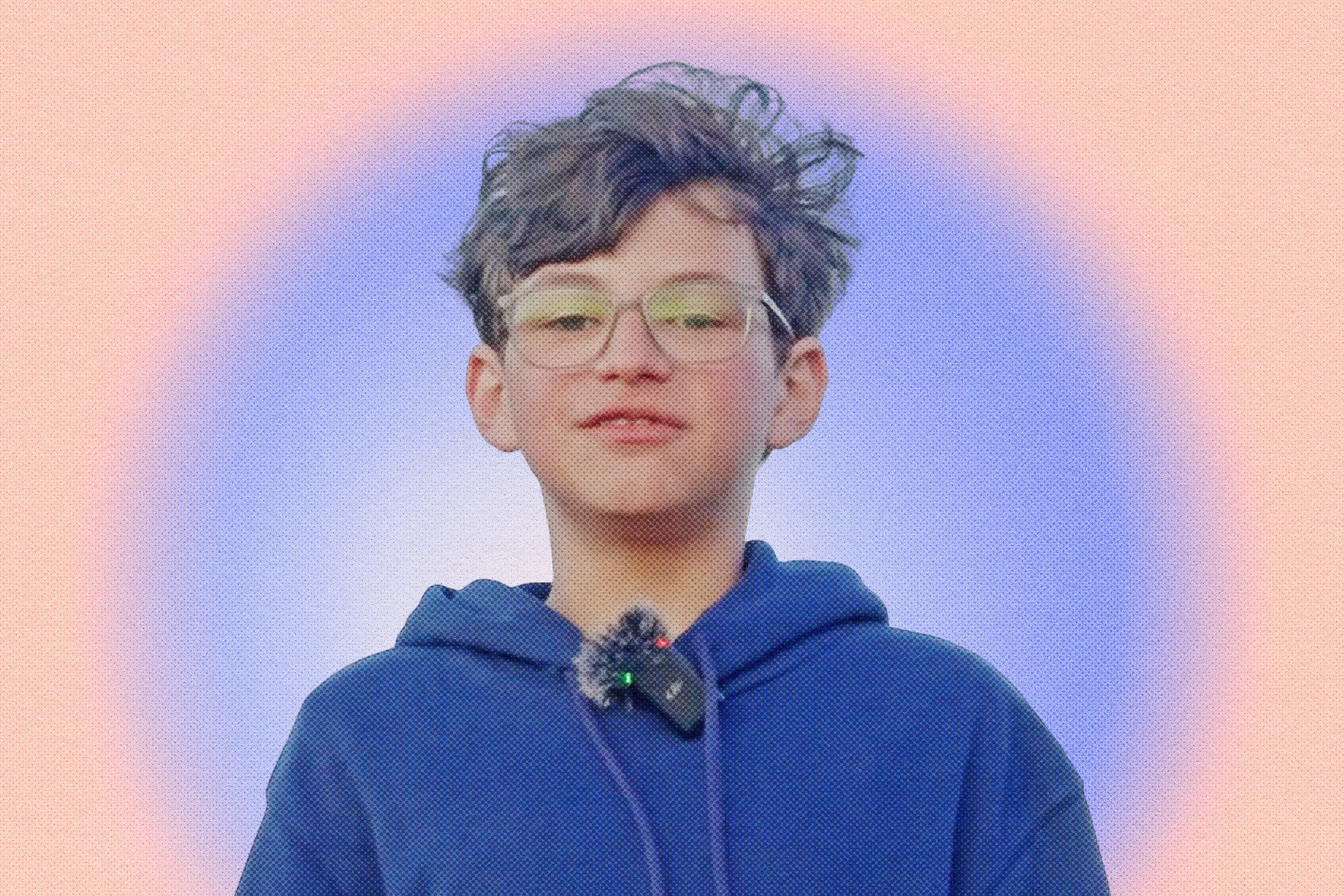 A young person with short, curly hair and glasses wears a blue hoodie with a microphone clipped on, against a soft purple and orange gradient background.
