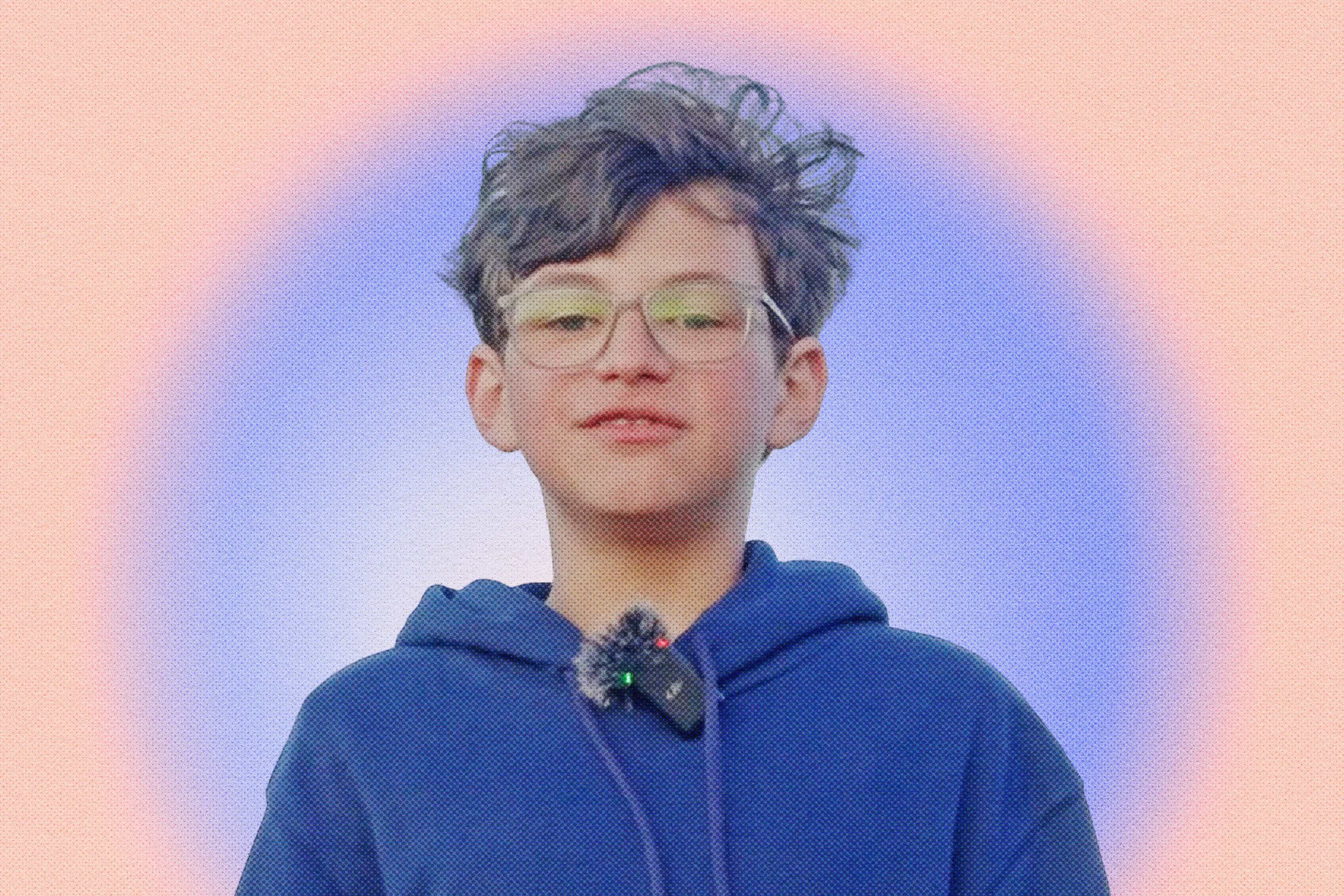 A young person with short, curly hair and glasses wears a blue hoodie with a microphone clipped on, against a soft purple and orange gradient background.