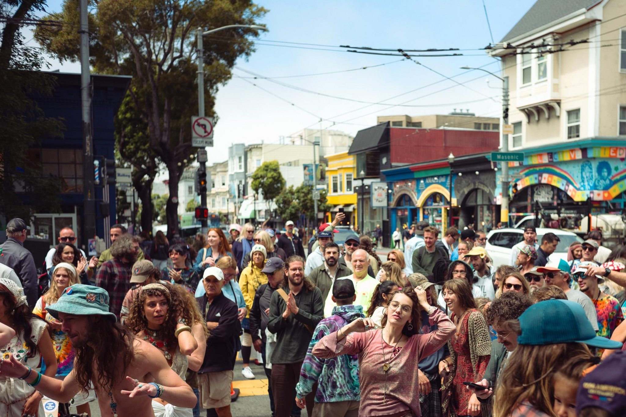 A diverse crowd of people happily dance and gather on a bright city street with colorful murals and buildings in the background.