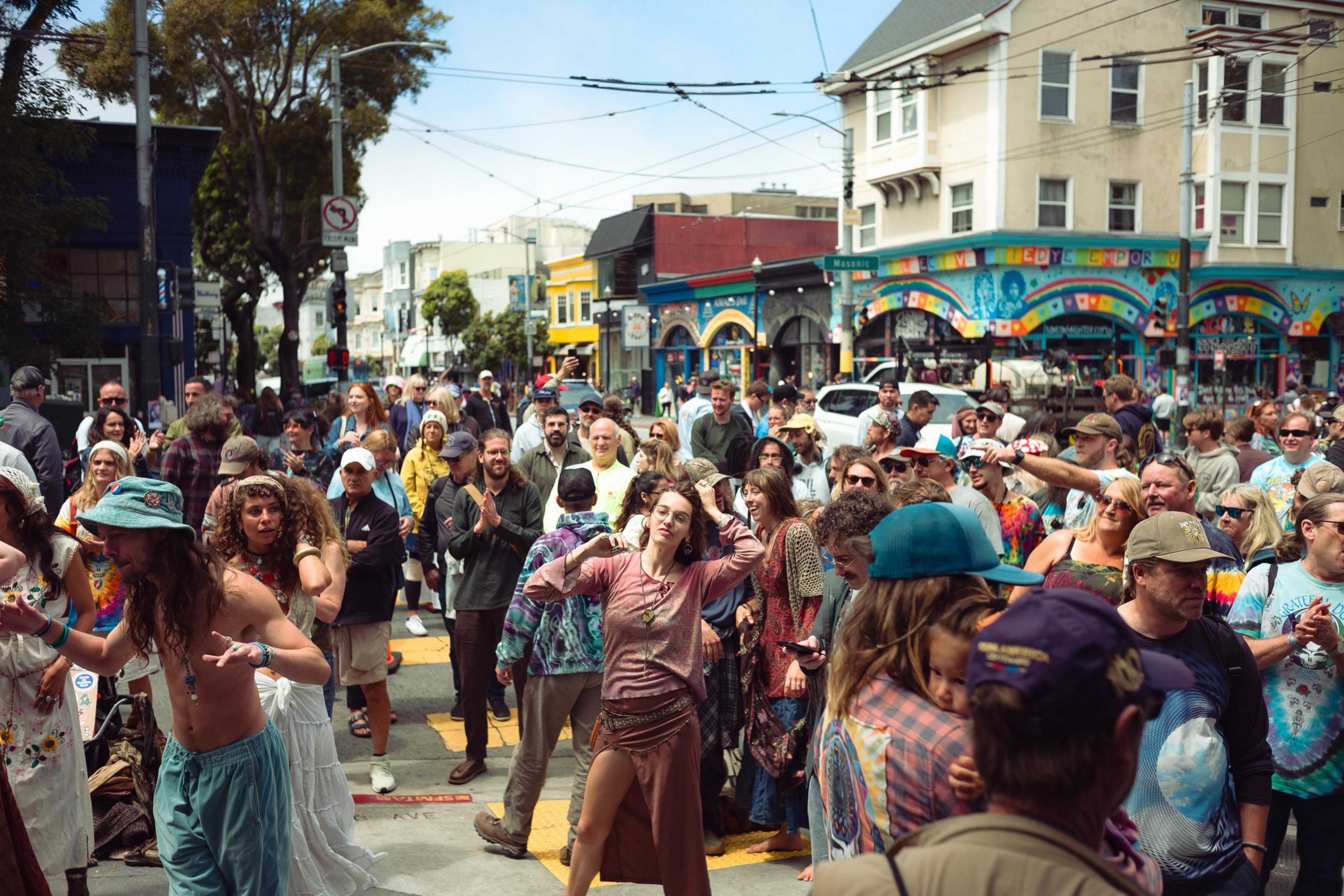 A diverse crowd of people happily dance and gather on a bright city street with colorful murals and buildings in the background.