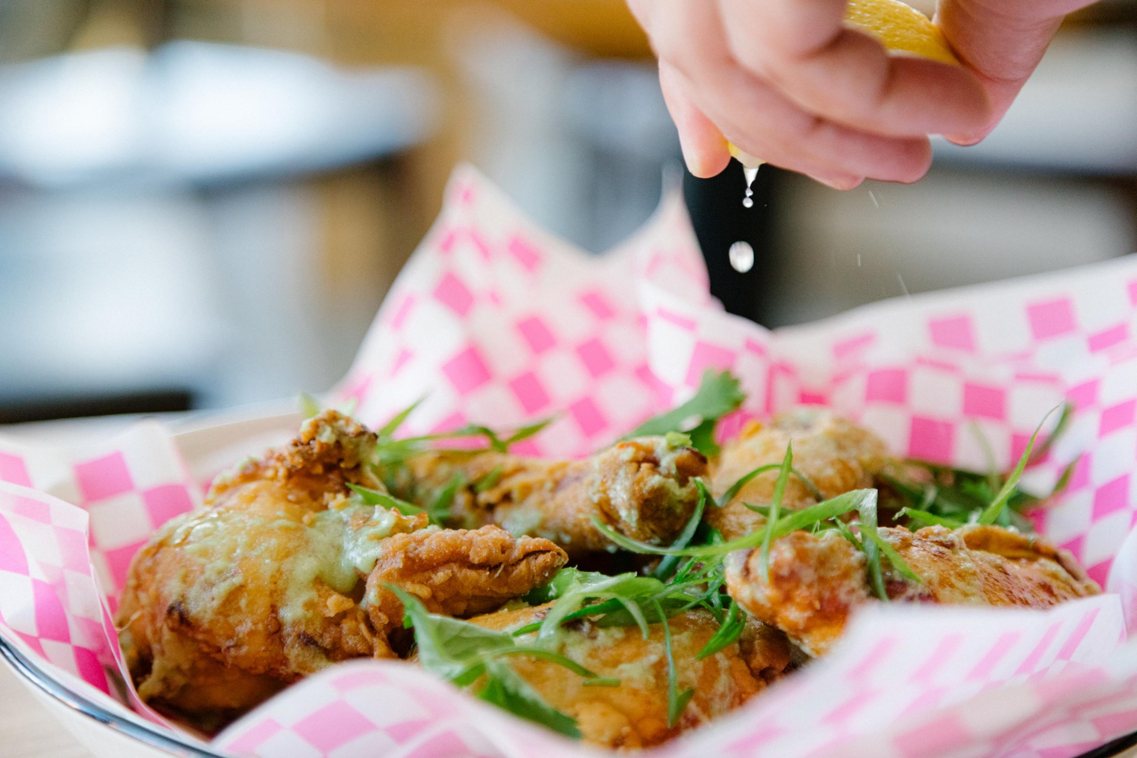 A hand squeezes lemon juice over crispy fried chicken garnished with fresh greens, set on pink checkered paper.