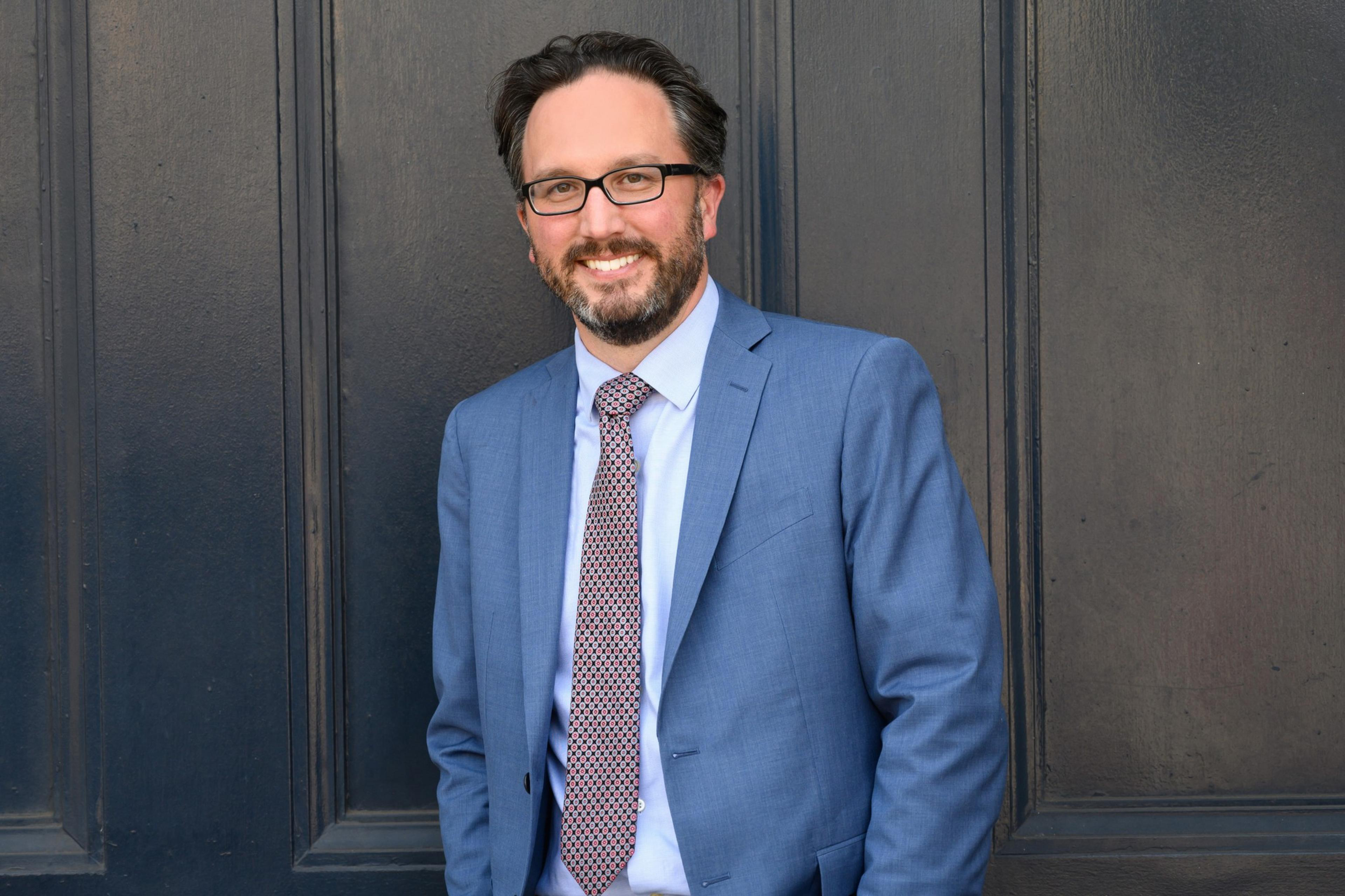 A man with short dark hair and glasses is smiling, wearing a blue suit, patterned tie, and white shirt, standing against a dark, textured wall.