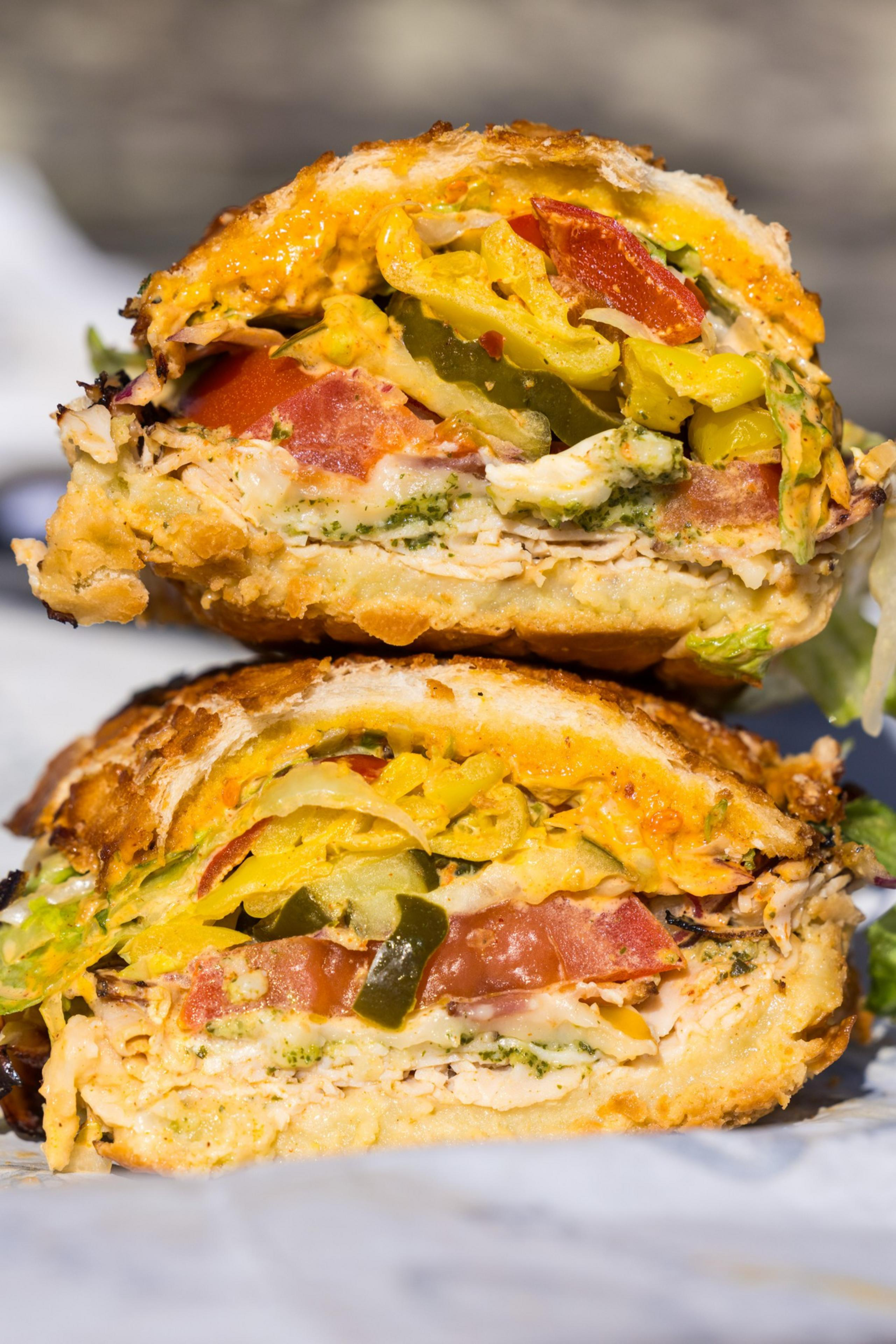 A sandwich cut in half, stacked with chicken, lettuce, tomatoes, cheddar, and pickles inside crusty bread, drizzled with a sauce.