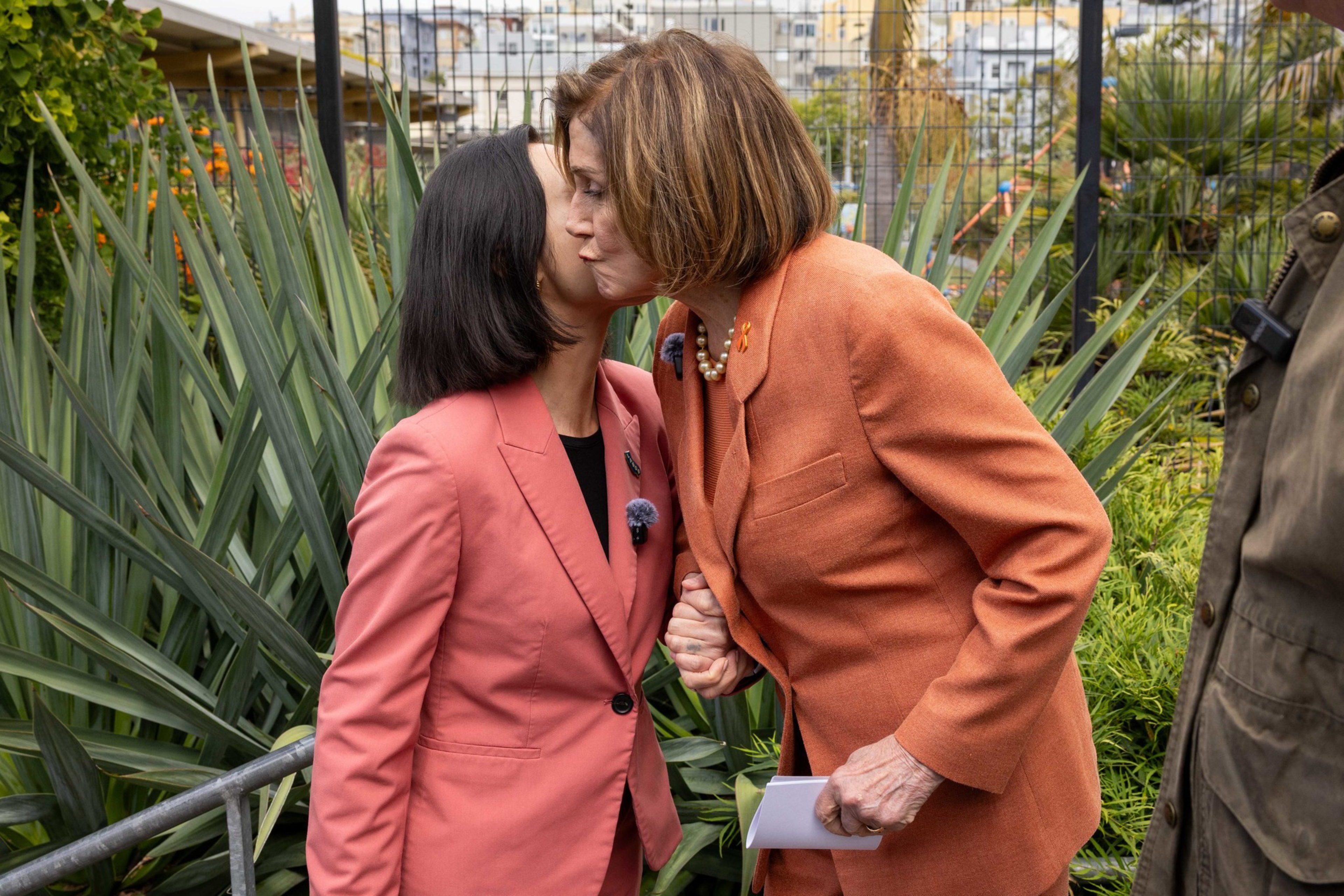 Two women in orange blazers greet each other with a cheek kiss and hold hands amid green plants in an outdoor garden setting.