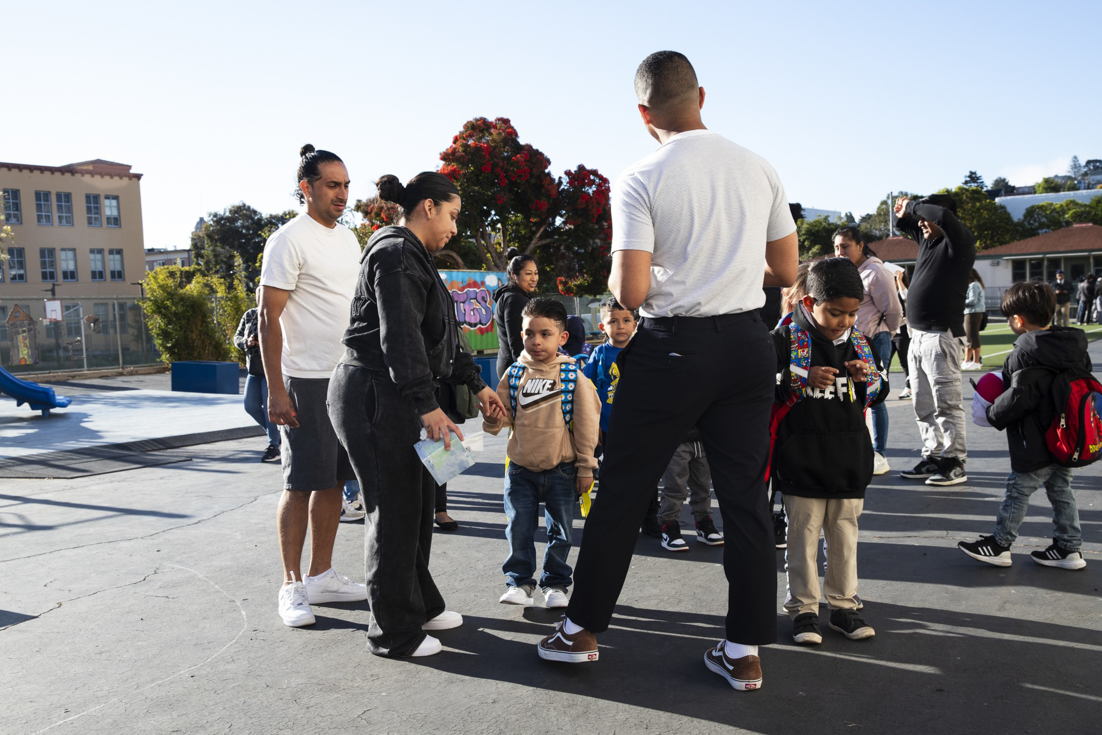 Parents and children gather outdoors on a sunny day, with some kids wearing backpacks and adults talking or helping them.