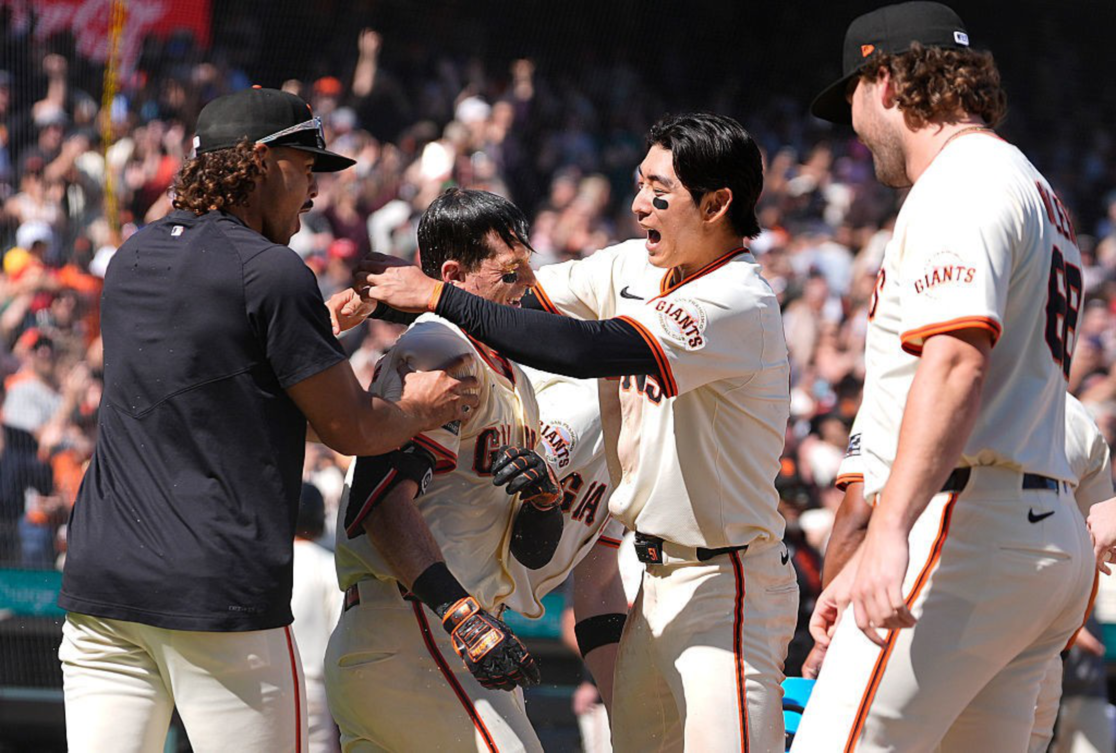 A group of baseball players in white Giants uniforms are celebrating energetically on the field, surrounded by cheering fans in the background.