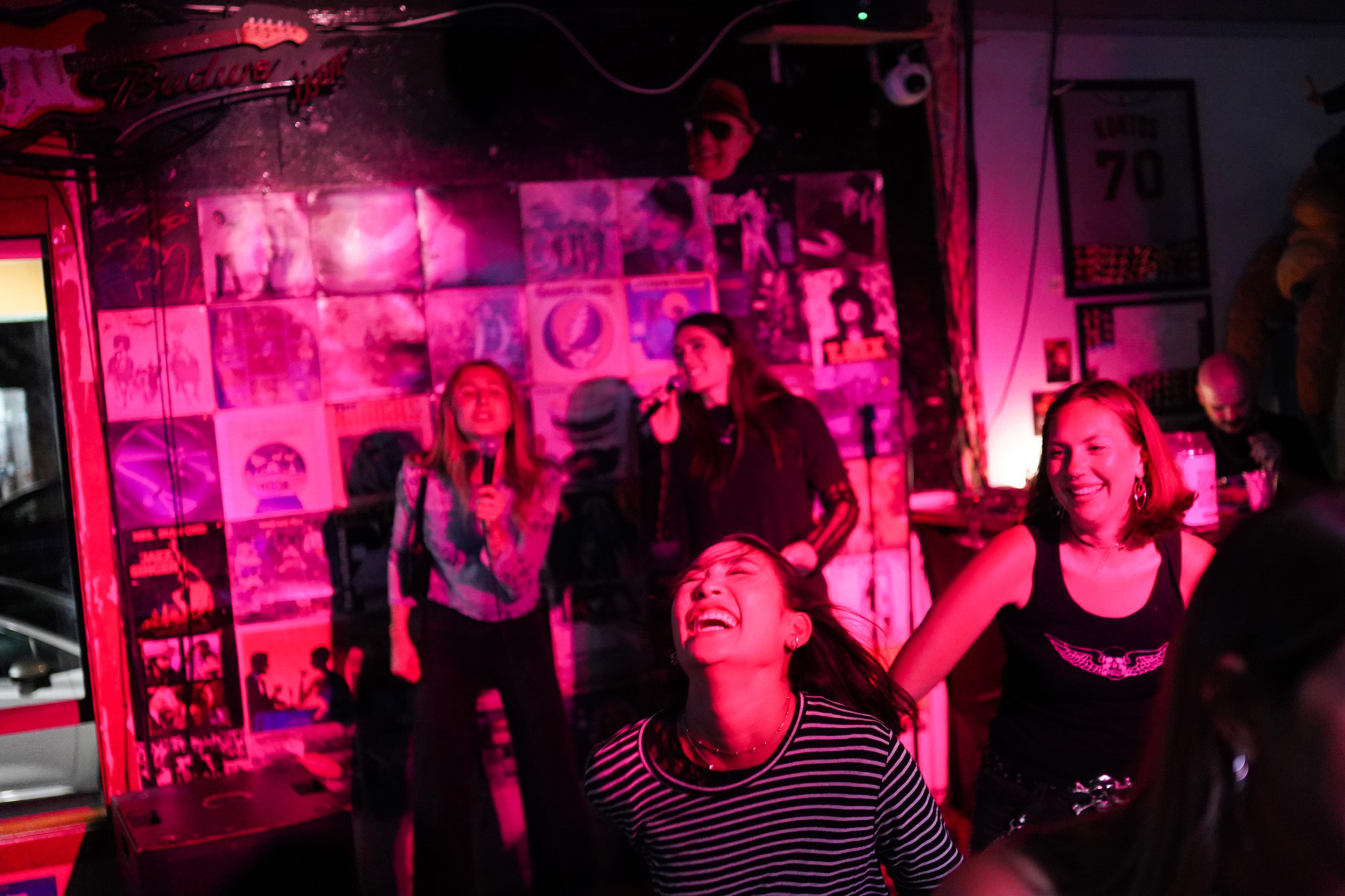 People sing karaoke on a colorful wall of album covers, while others dance and smile under vibrant pink and purple lighting.