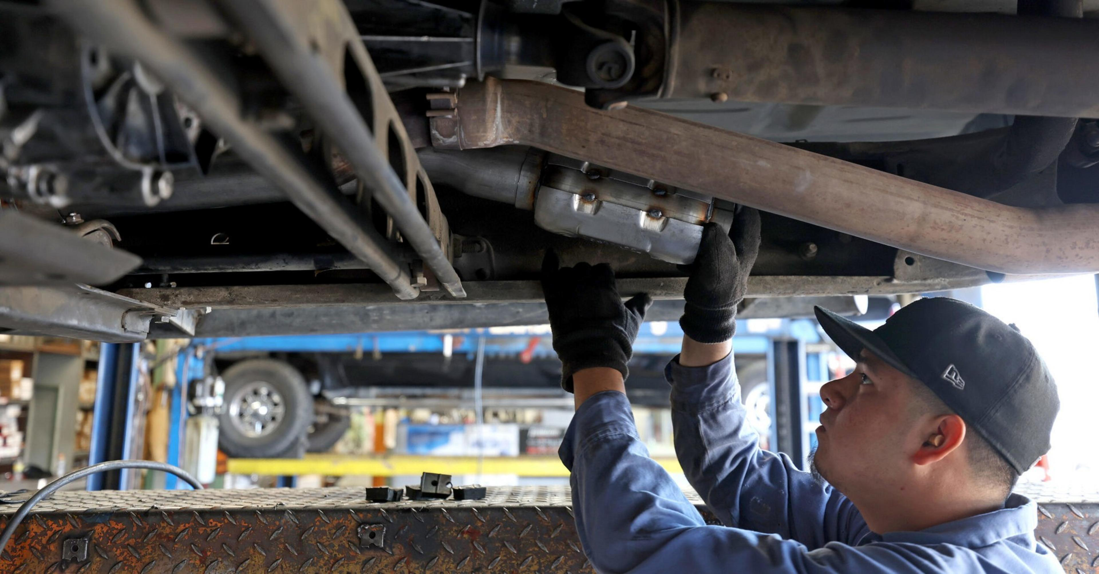 A mechanic wearing gloves and a cap works under a raised vehicle, inspecting or repairing parts underneath.