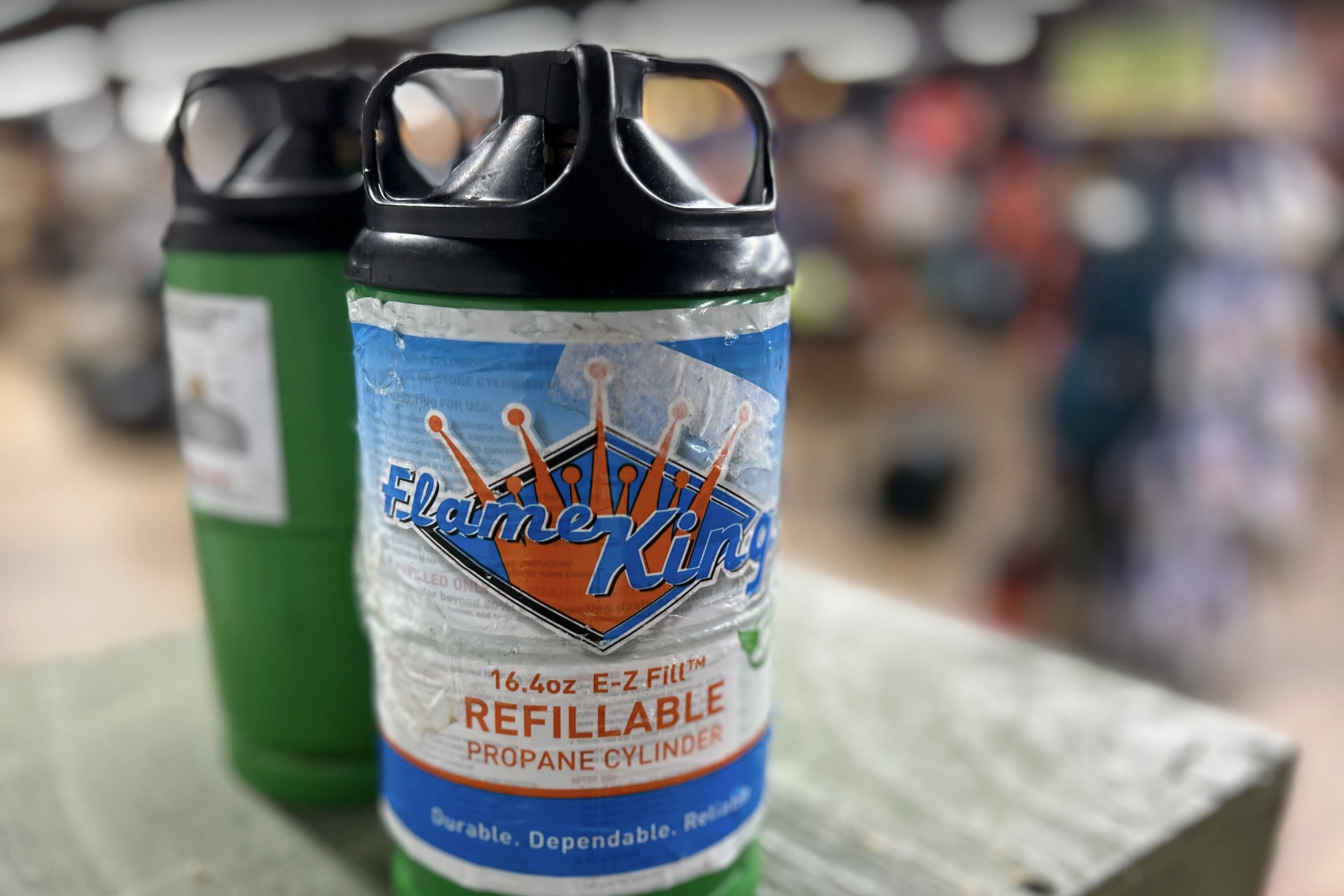 The image shows two green Flame King refillable propane cylinders with black caps, standing close together on a wooden surface in a blurred indoor setting.