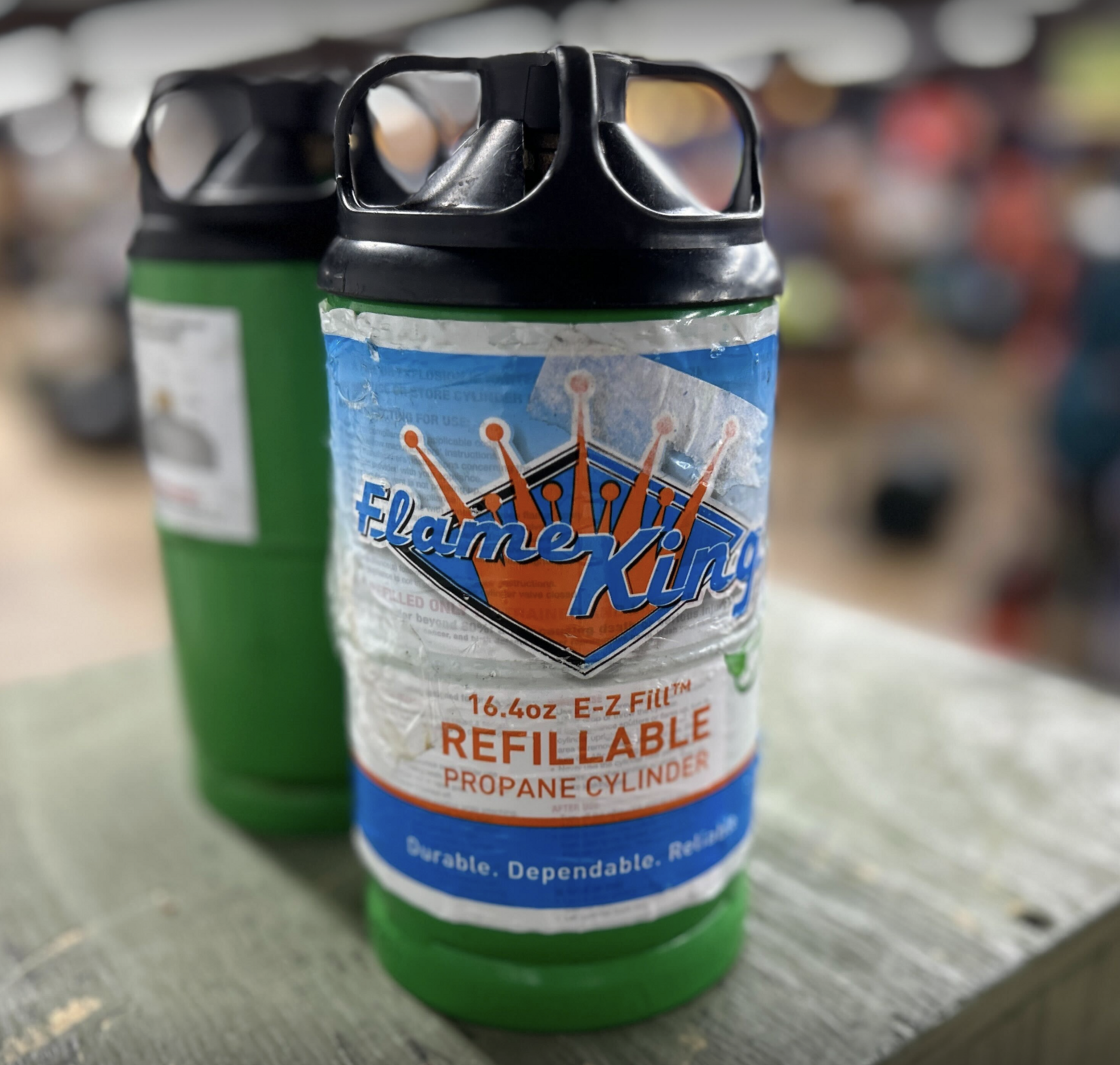 The image shows two green Flame King refillable propane cylinders with black caps, standing close together on a wooden surface in a blurred indoor setting.