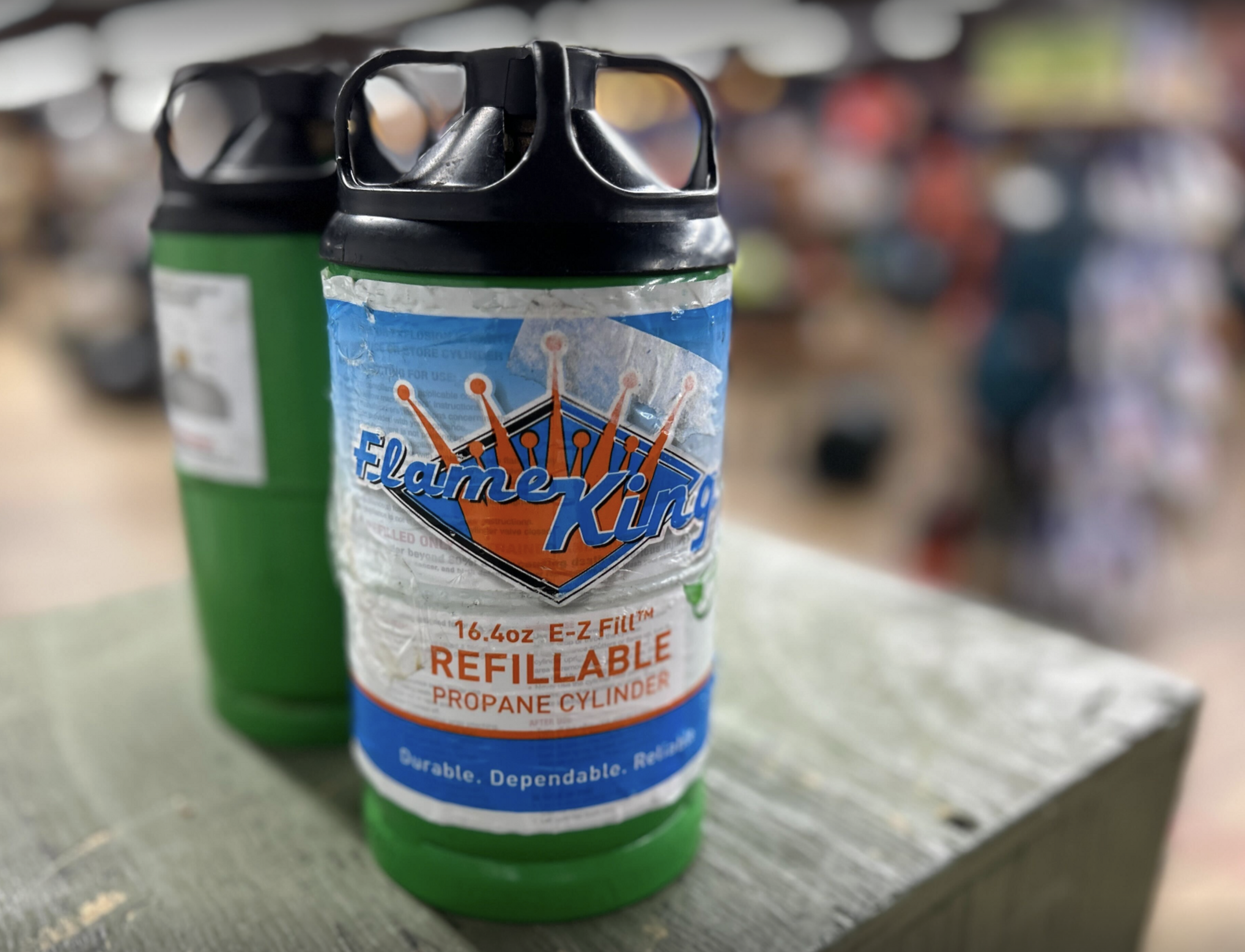The image shows two green Flame King refillable propane cylinders with black caps, standing close together on a wooden surface in a blurred indoor setting.