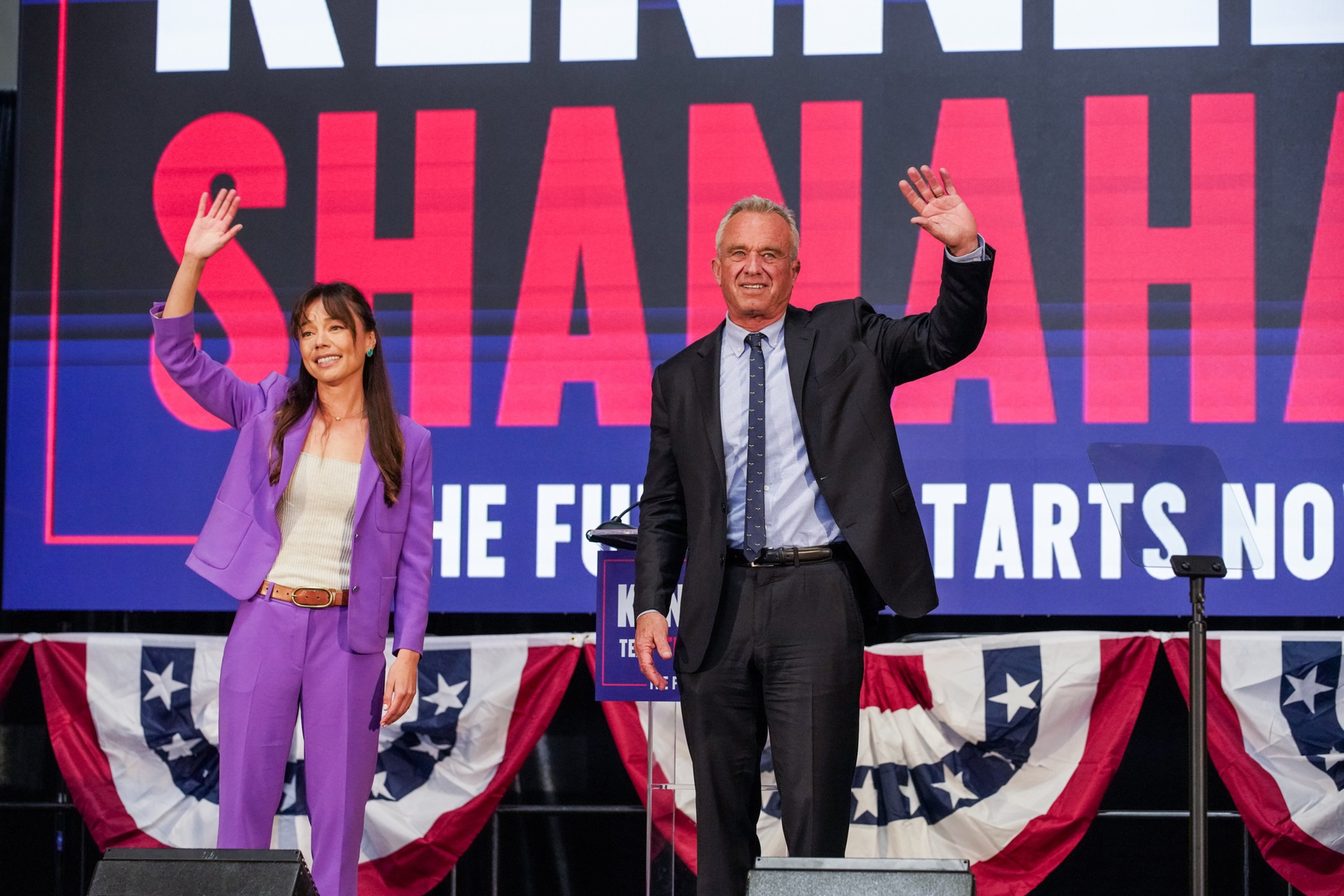 A man and woman wave to a crowd, standing before a banner with patriotic decor and a slogan about the future.