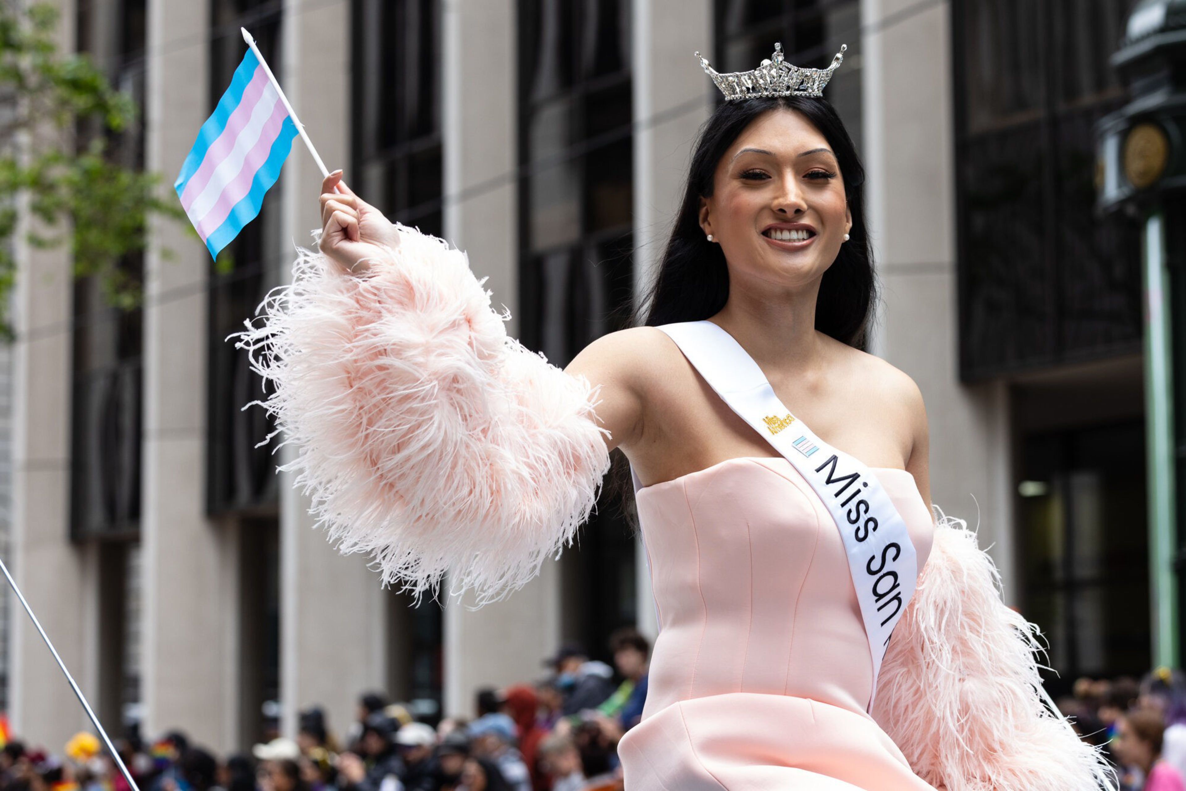 A crowned person wearing a pink dress and sash reading "Miss Son" waves a transgender pride flag during an outdoor event.