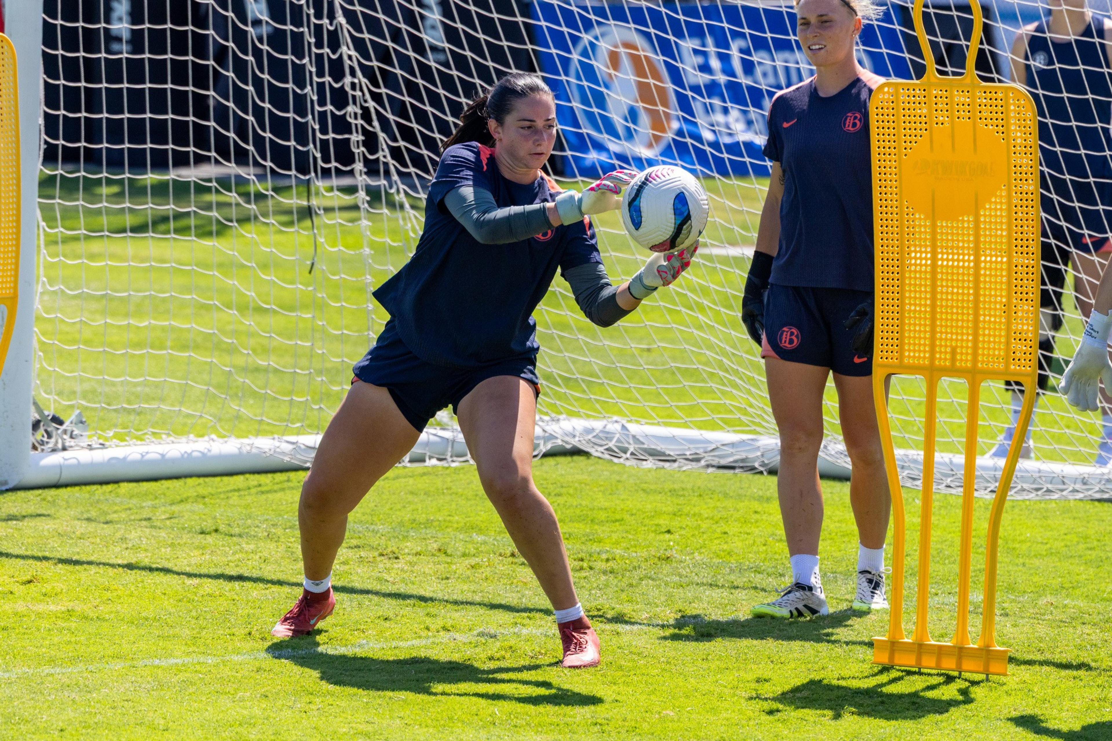 A female goalkeeper in dark athletic gear catches a soccer ball near the goal while another player and a training dummy stand nearby on the grass.