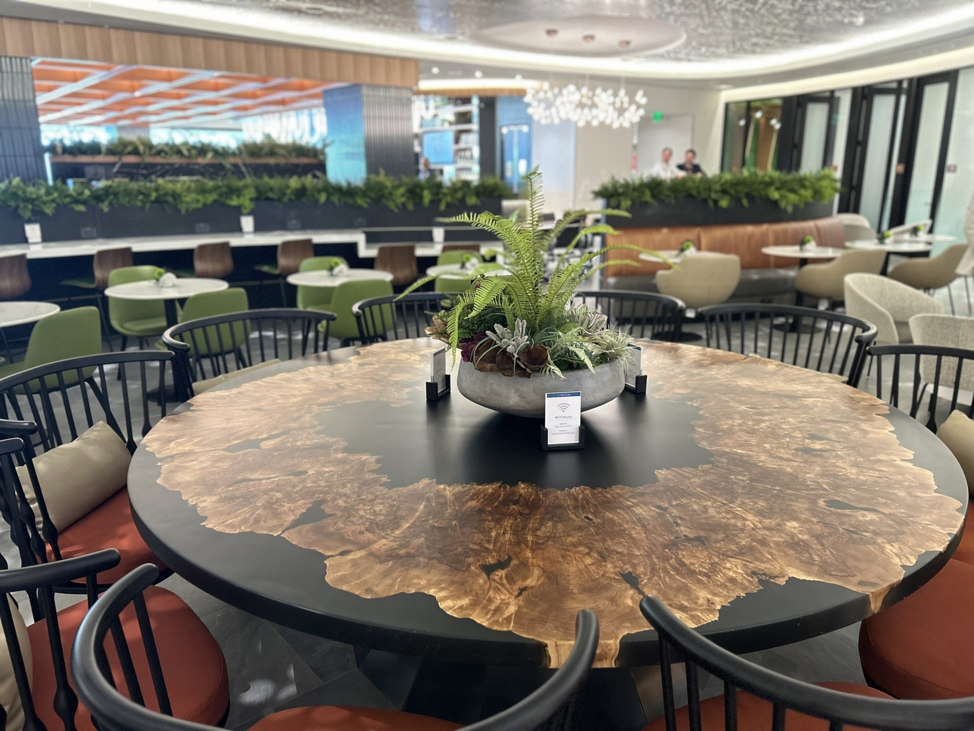 The image shows a modern cafe with a round table featuring a polished wood and black design, surrounded by black chairs with red cushions, and a plant centerpiece.