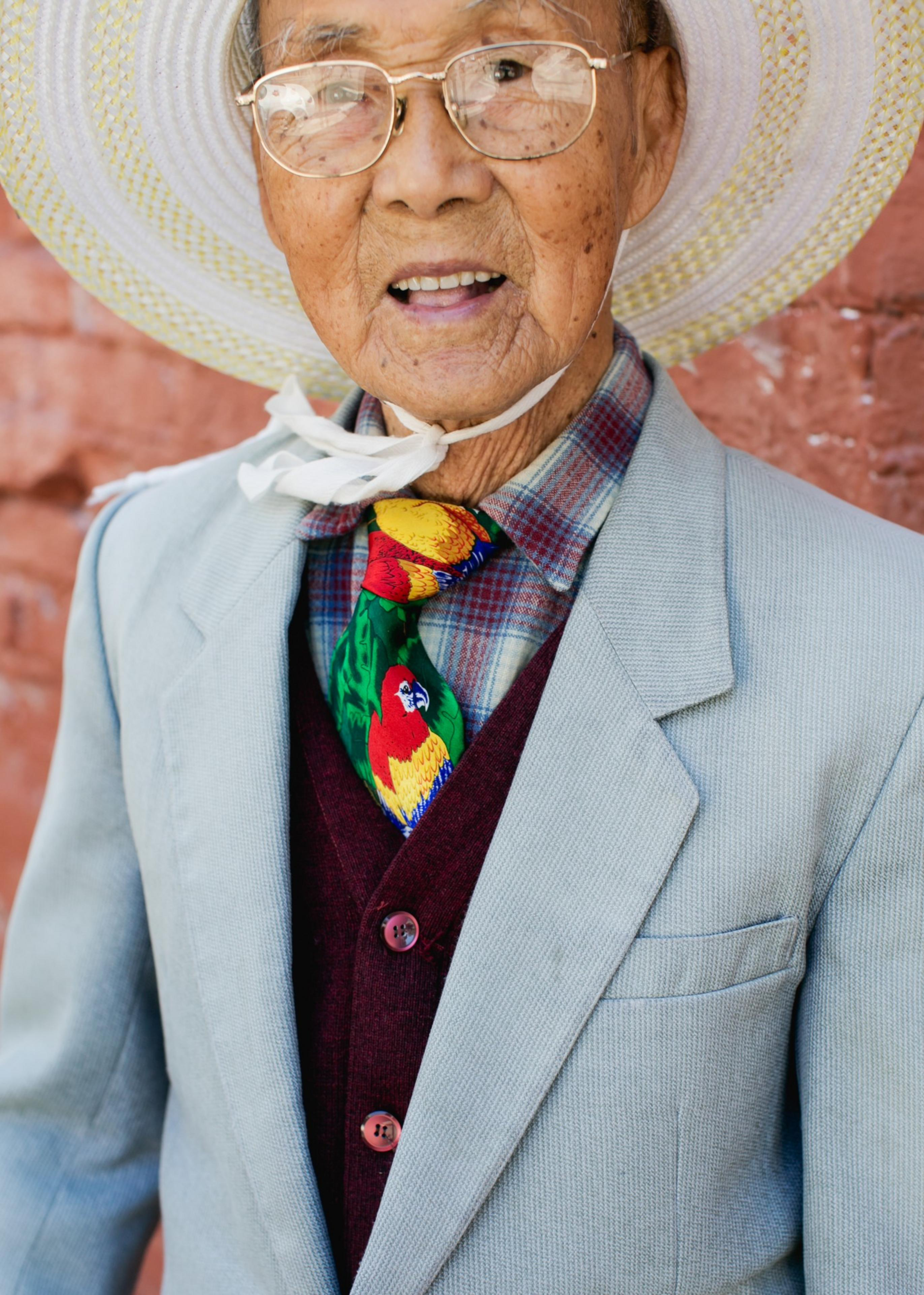 An elderly man wears a white wide-brimmed hat, glasses, a grey suit jacket, a burgundy vest, a plaid shirt, and a colorful parrot-patterned tie.