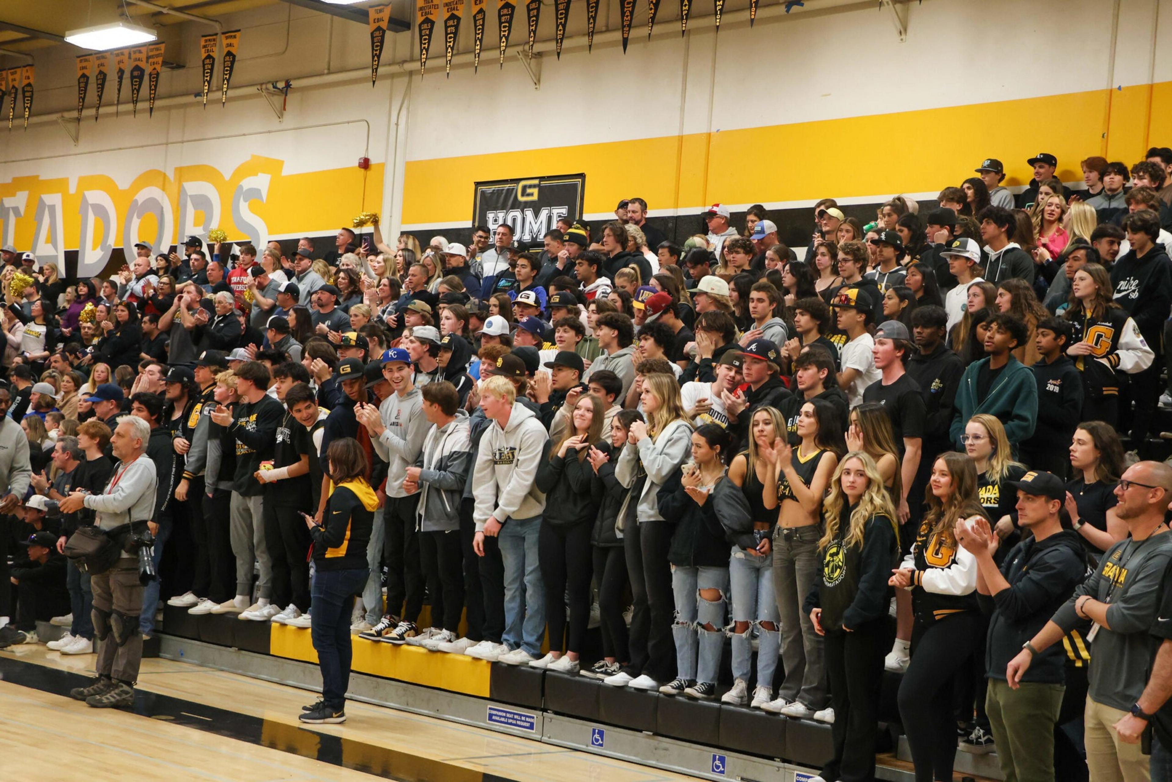 A large crowd of students and adults stand and cheer inside a gym with yellow and black walls and "MATADORS" written on the wall.
