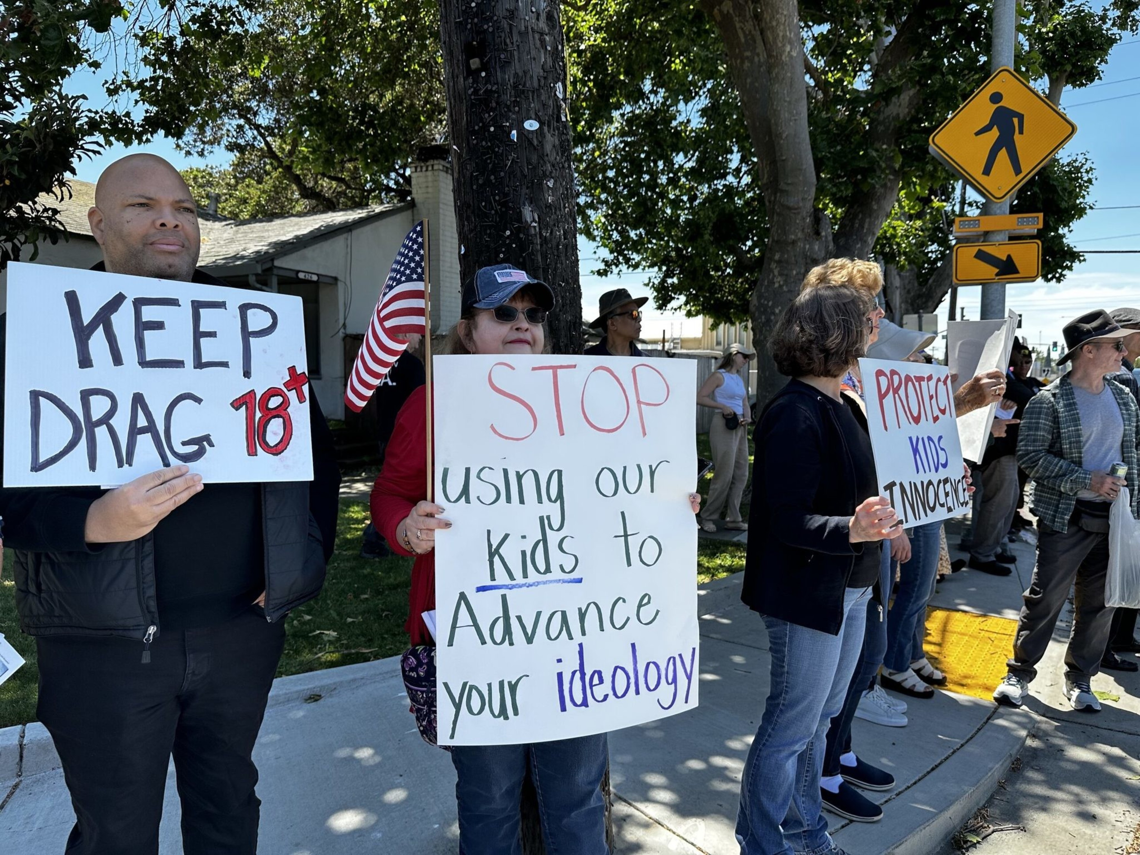 A group of people stand on a sidewalk holding signs reading "KEEP DRAG 18+," "STOP using our kids to Advance your ideology," and "PROTECT KIDS INNOCENCE."
