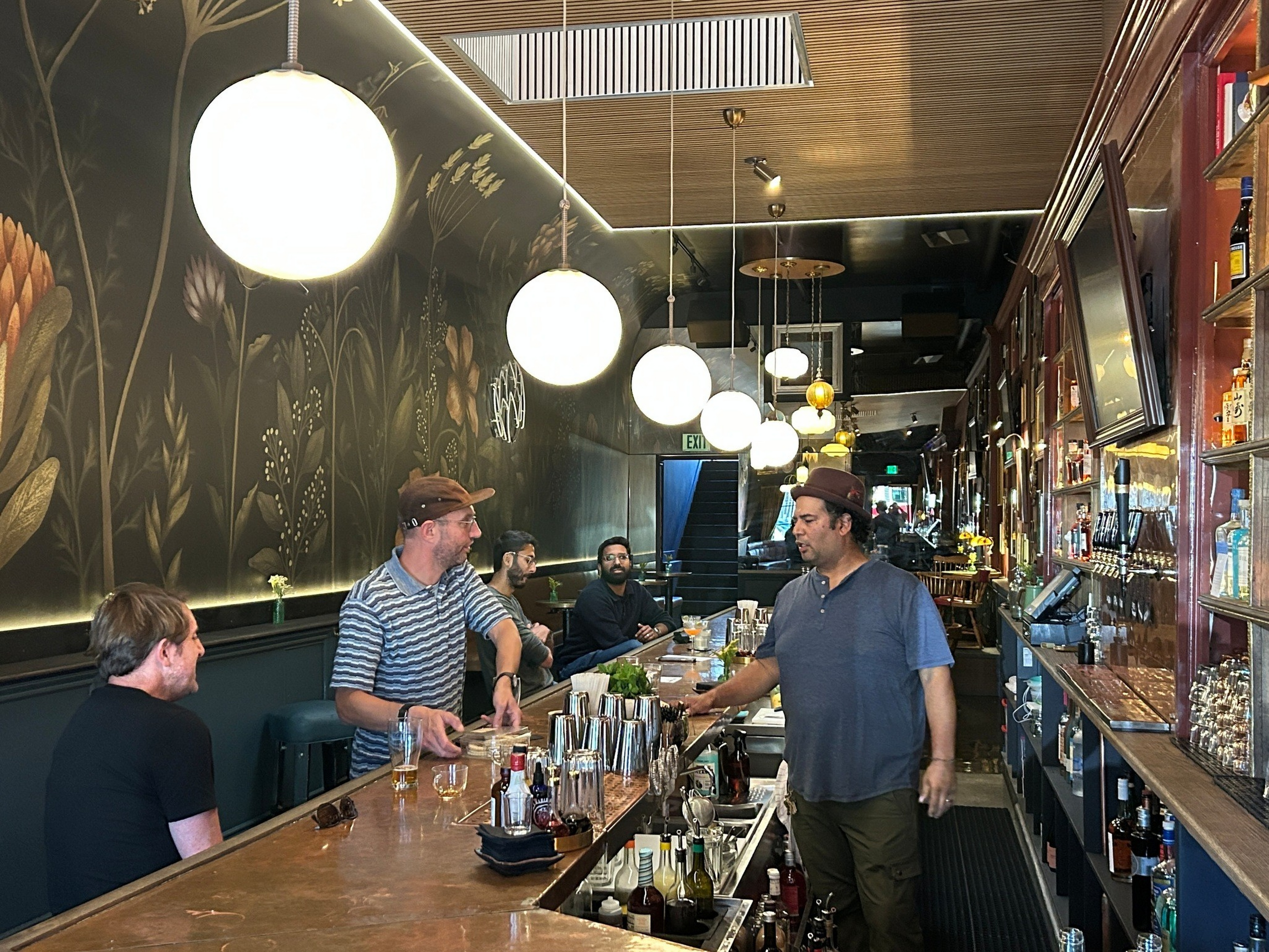 A cozy bar with floral wallpaper, warm hanging lights, a bartender talking to customers seated at the counter with drinks and bar tools around.