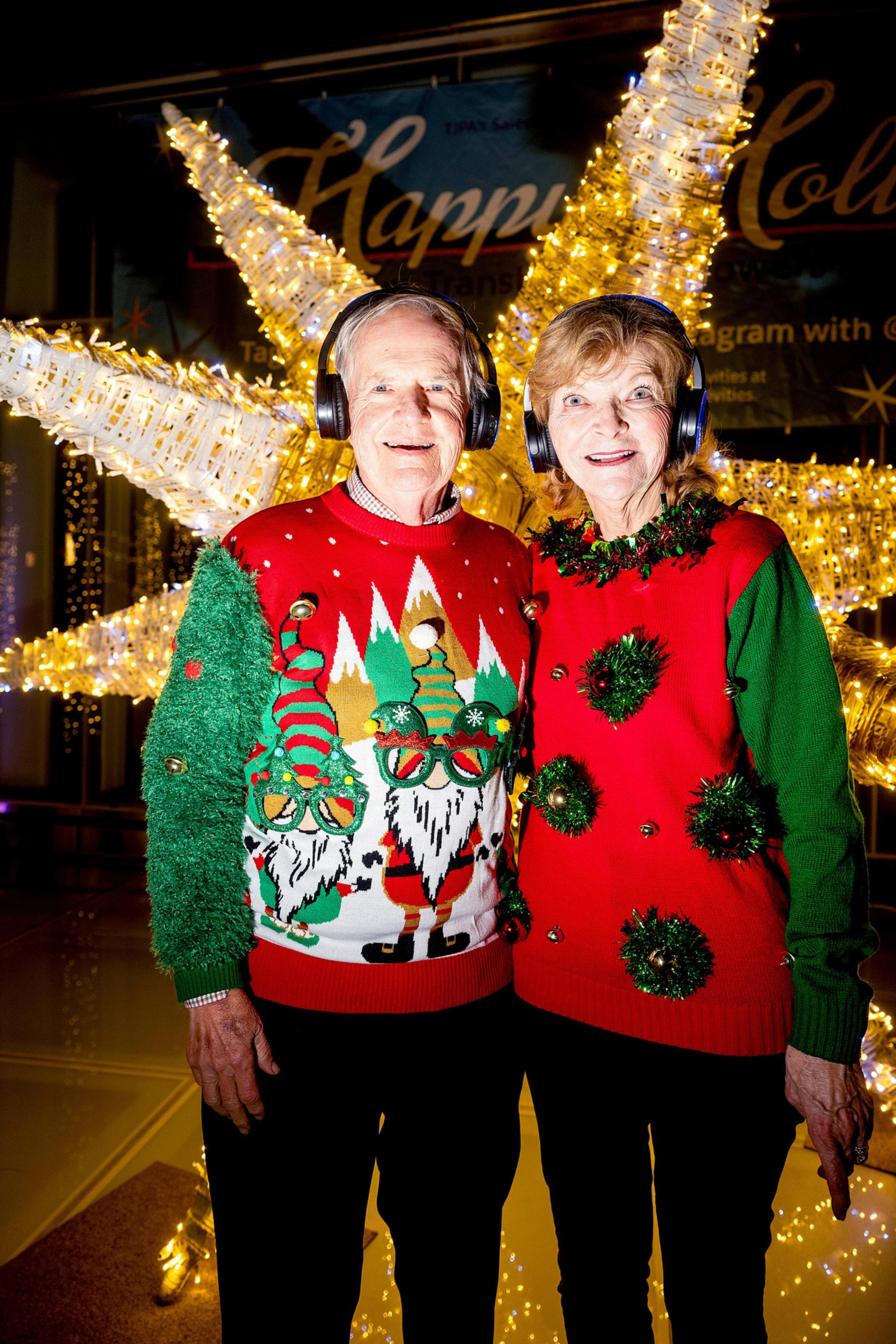 An elderly couple in festive sweaters and headphones smiles in front of a large, illuminated star. The background reads &quot;Happy Holidays&quot; with twinkling lights.