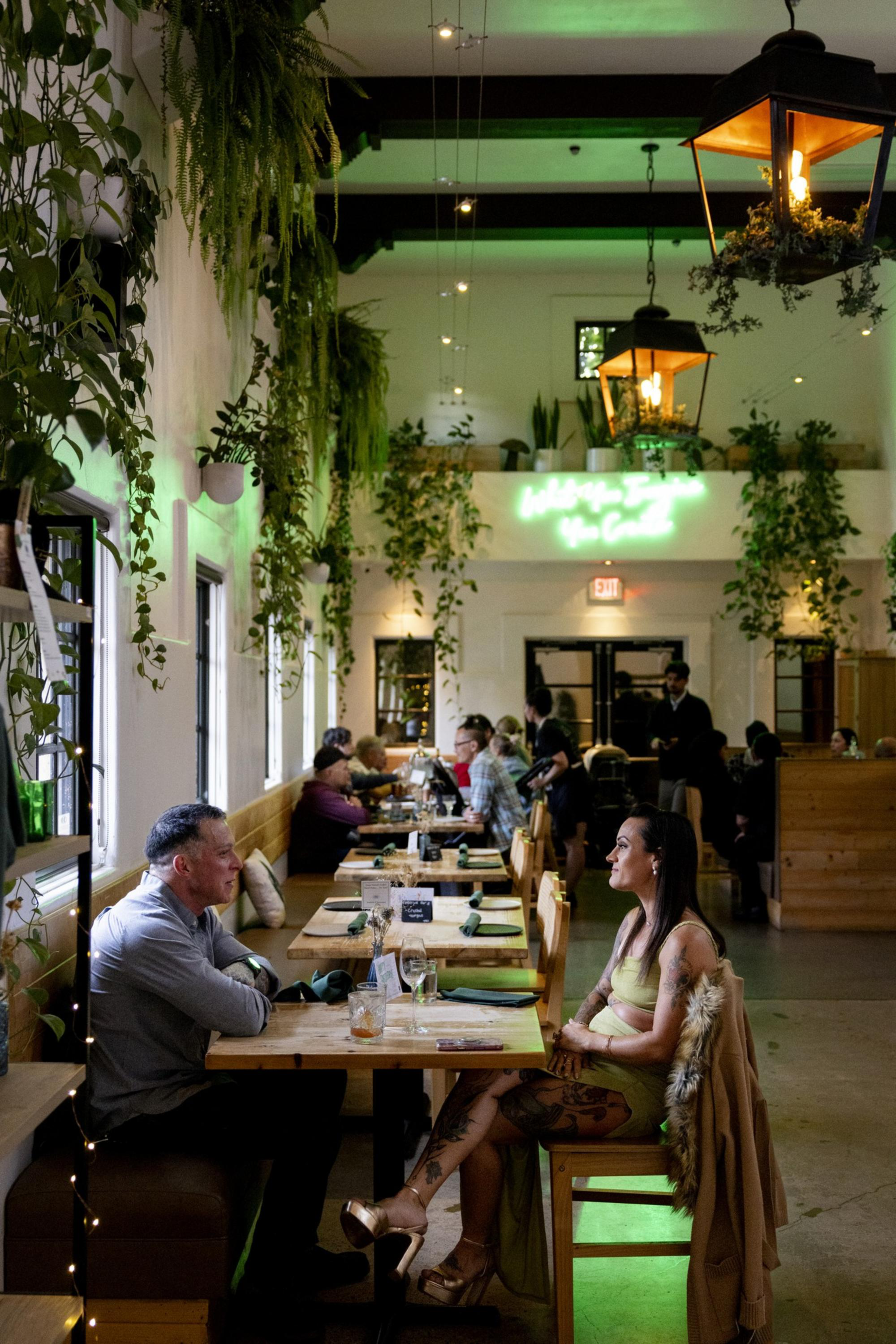 People sit at wooden tables in a lush, plant-filled restaurant with hanging greenery, warm lanterns, and a green neon sign on the back wall.
