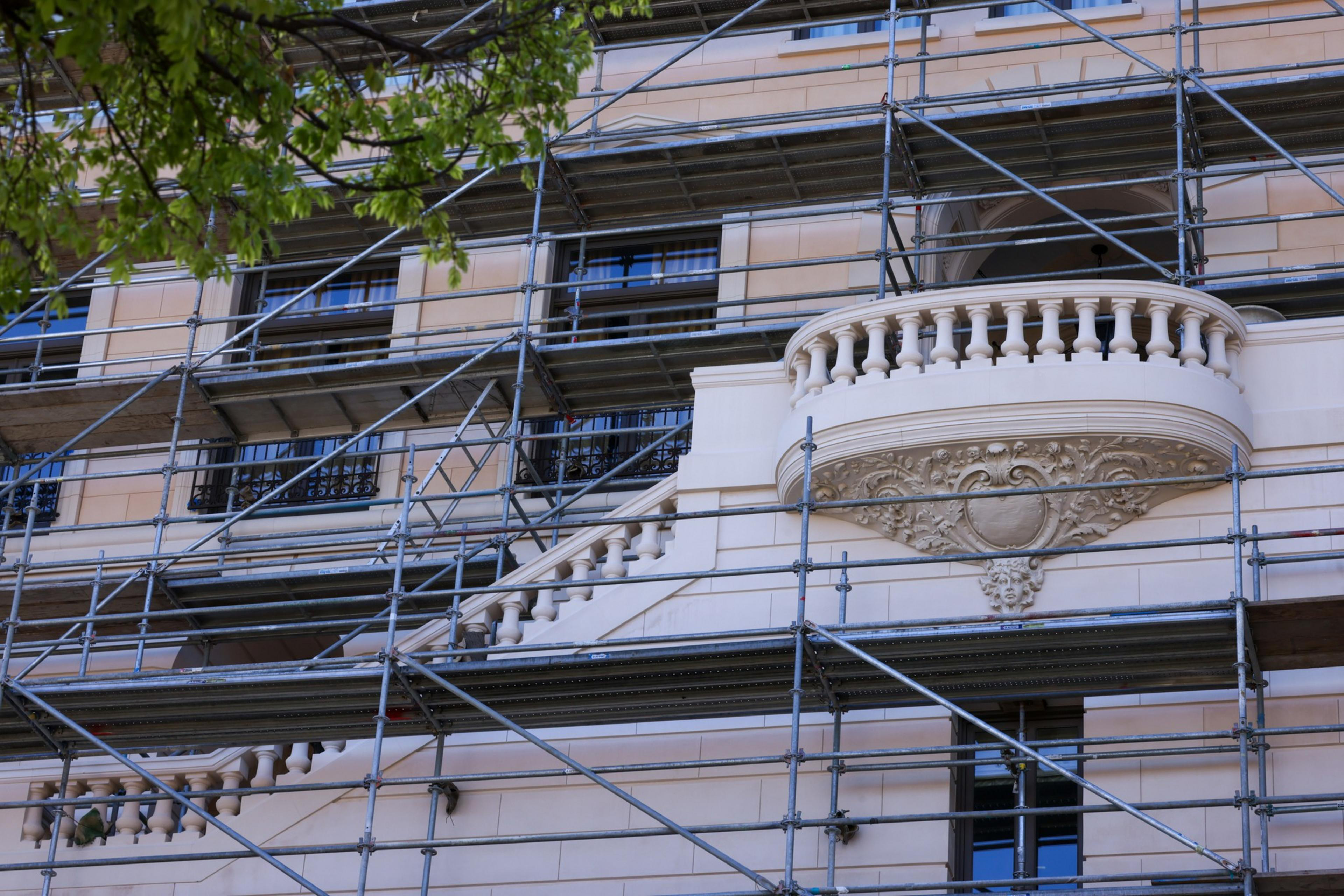 A building facade covered with scaffolding is shown, featuring intricate stonework and a small balcony. Leaves from a tree partially frame the top left.