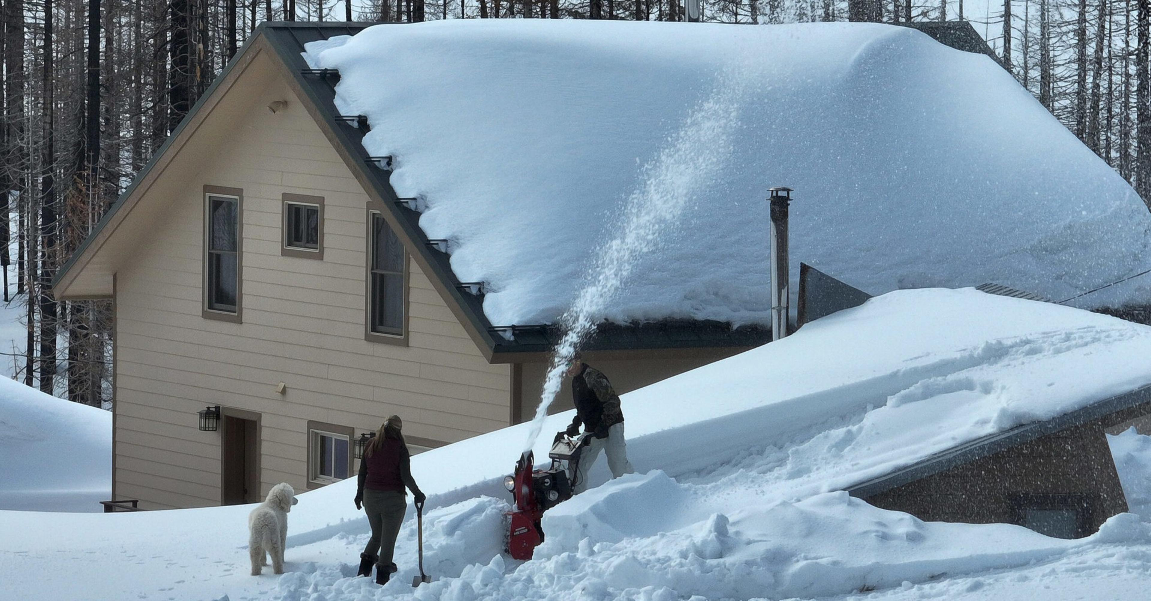 Two people clear deep snow with a snow blower and shovel near a house and garage, while a dog stands in the snow watching.