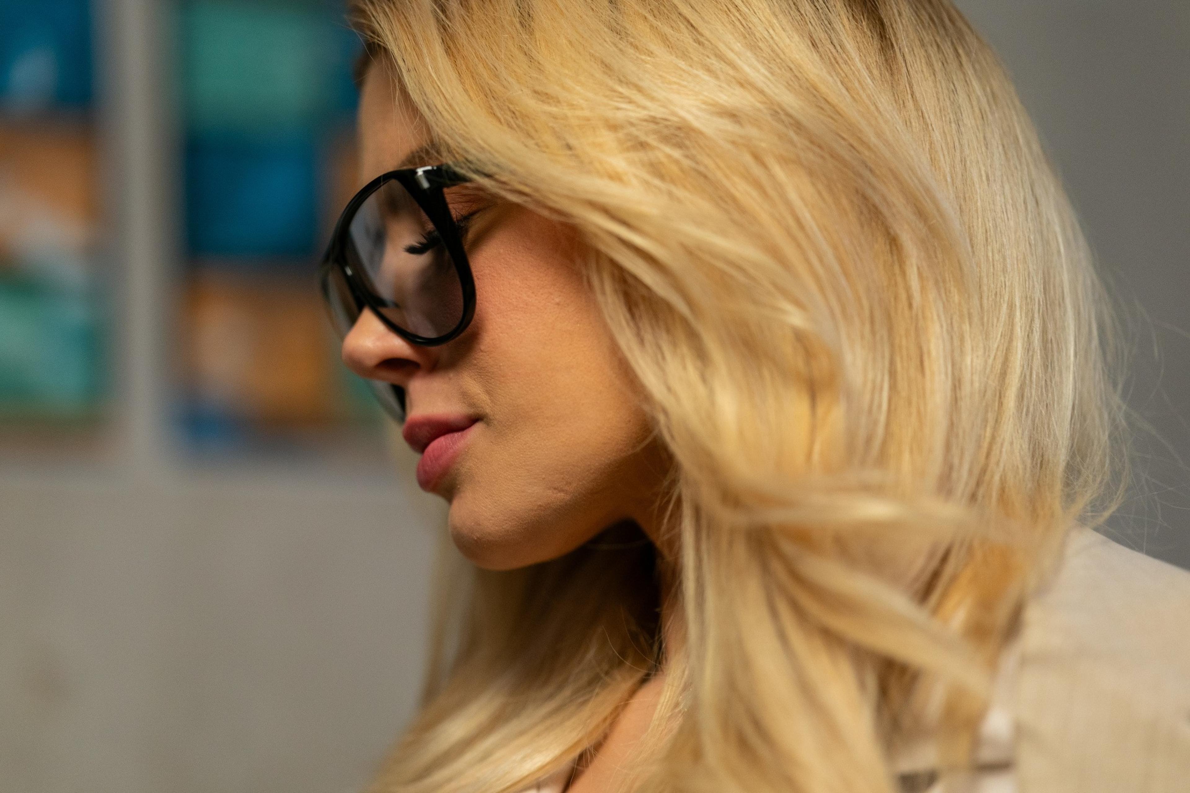 A close-up of a person with long blonde hair wearing large black glasses, facing sideways. The background is soft-focused with bluish and orange tones.
