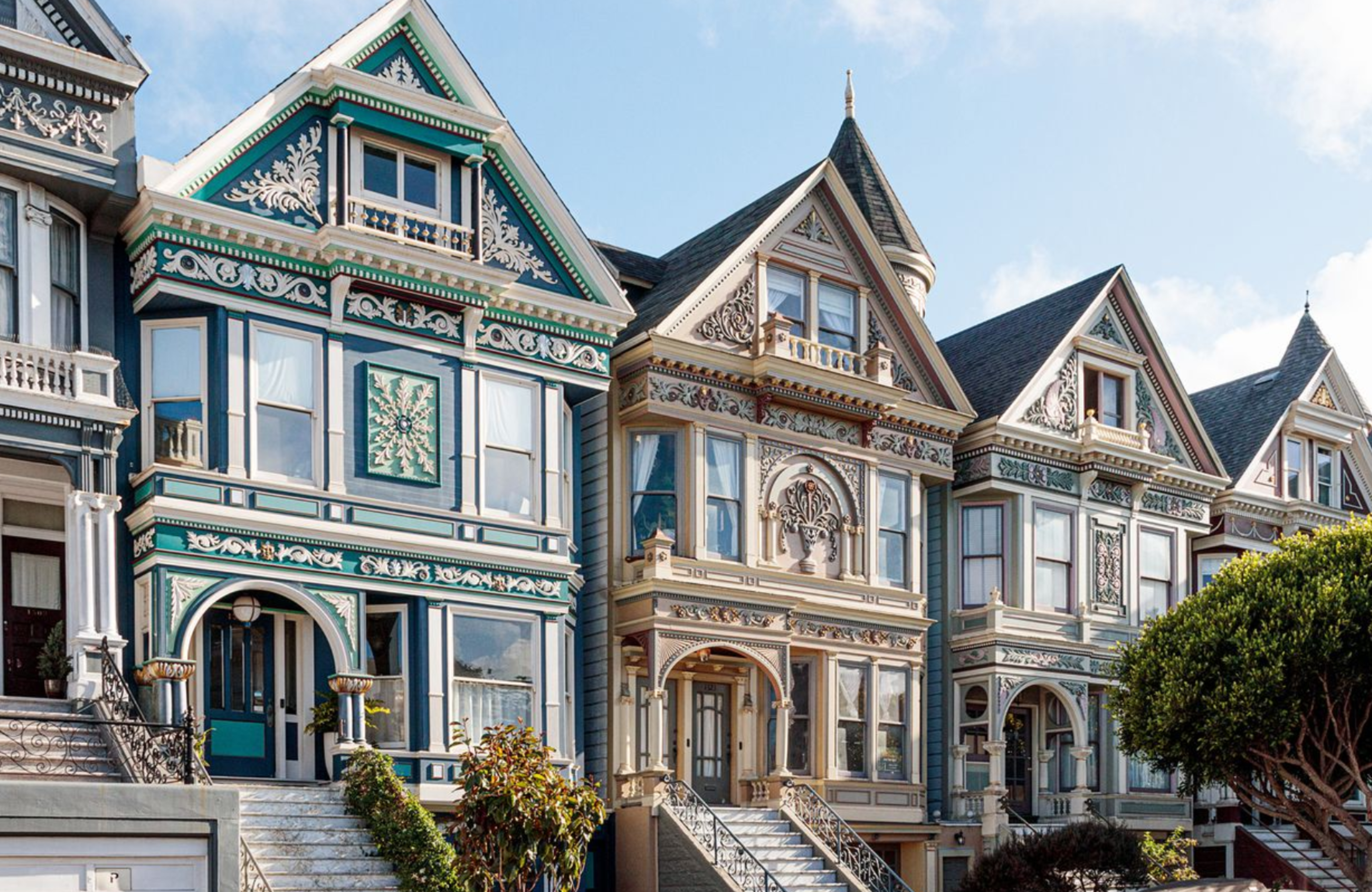 Four brightly colored Victorian-style houses with intricate woodwork, tall windows, and steep gabled roofs stand side by side under a clear sky.