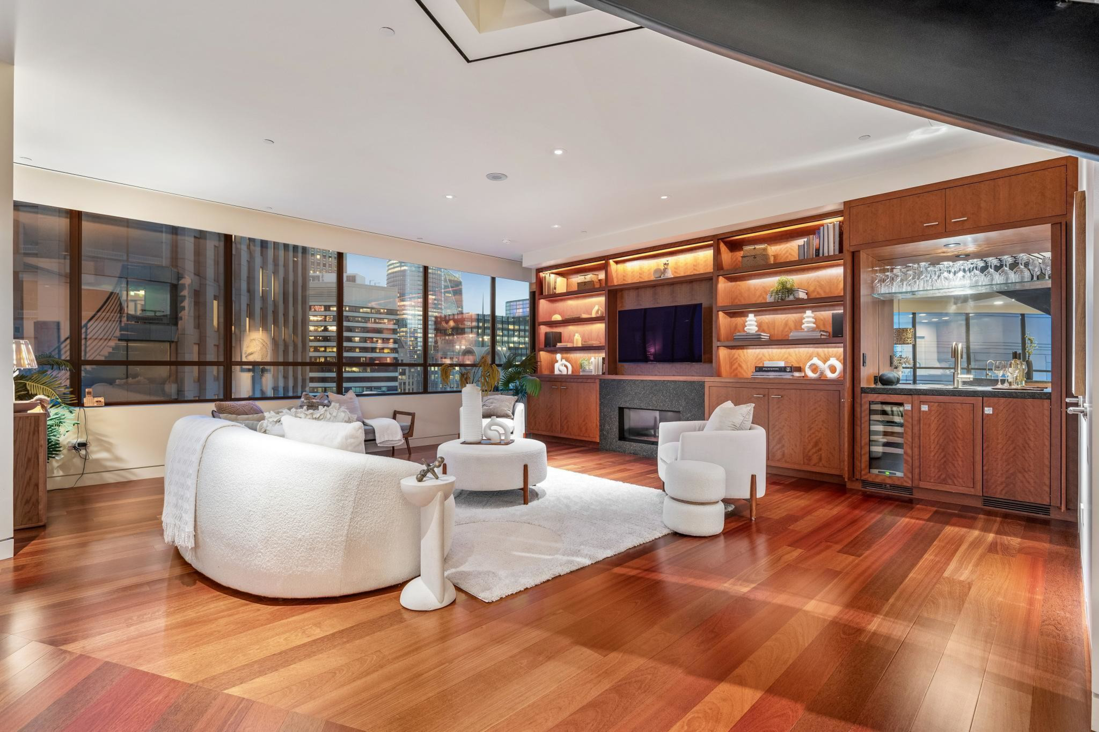 A modern living room with curved white sofa, round ottoman, wooden floor, built-in wooden shelves, fireplace, TV, and cityscape visible through large windows.